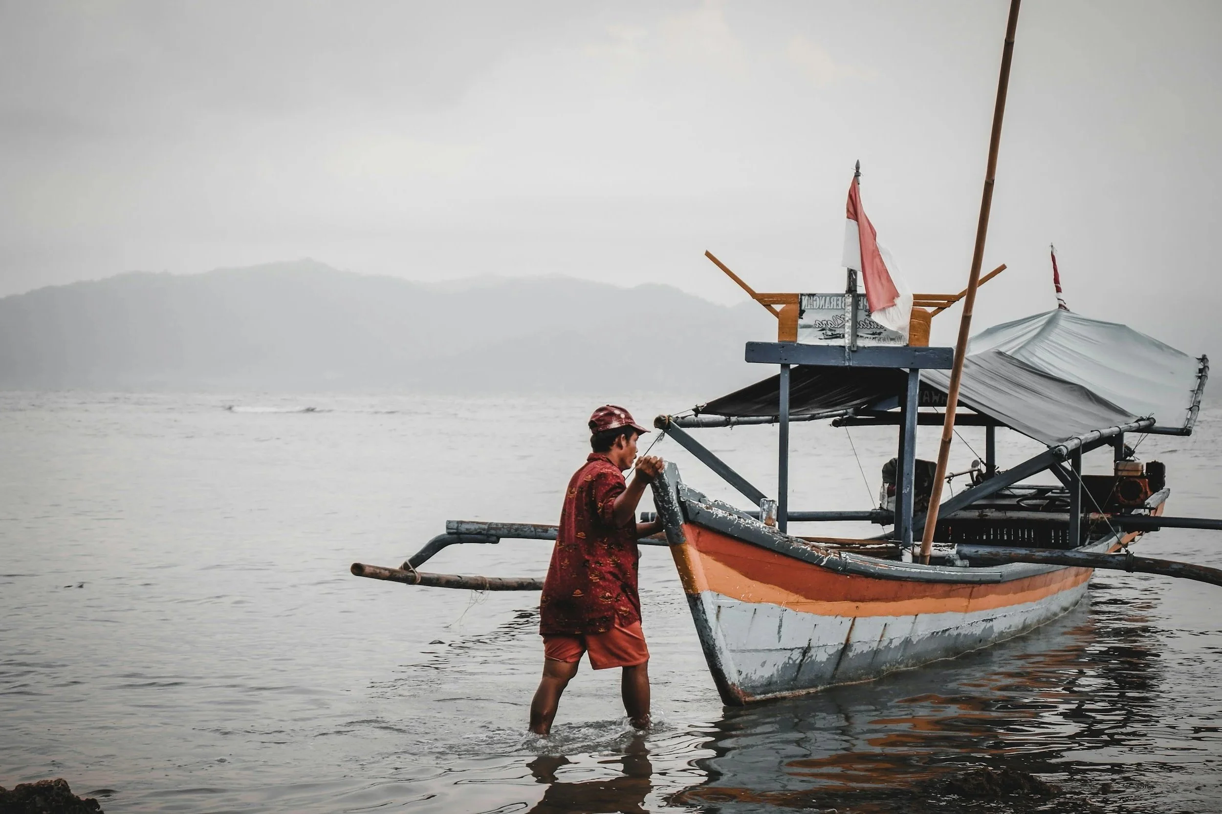 A person wearing a red patterned shirt and red shorts is standing in shallow water, holding onto a boat with a white and orange hull that is being floated near a rocky shore. The boat has a small wooden platform and a mast with a red and white flag, and there are hills or mountains in the background under an overcast sky.
