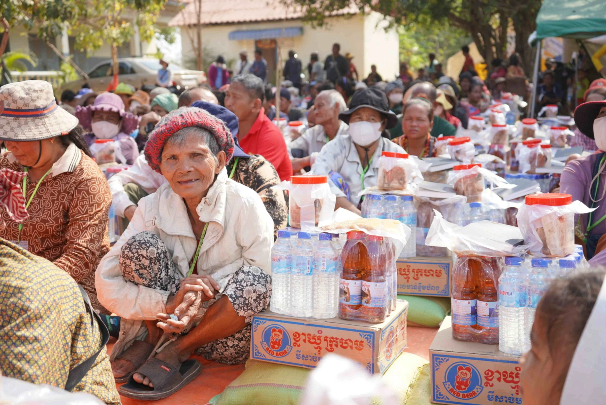 Elderly woman wearing a knit hat is sitting in crowded area with humanitarian supplies including food and water