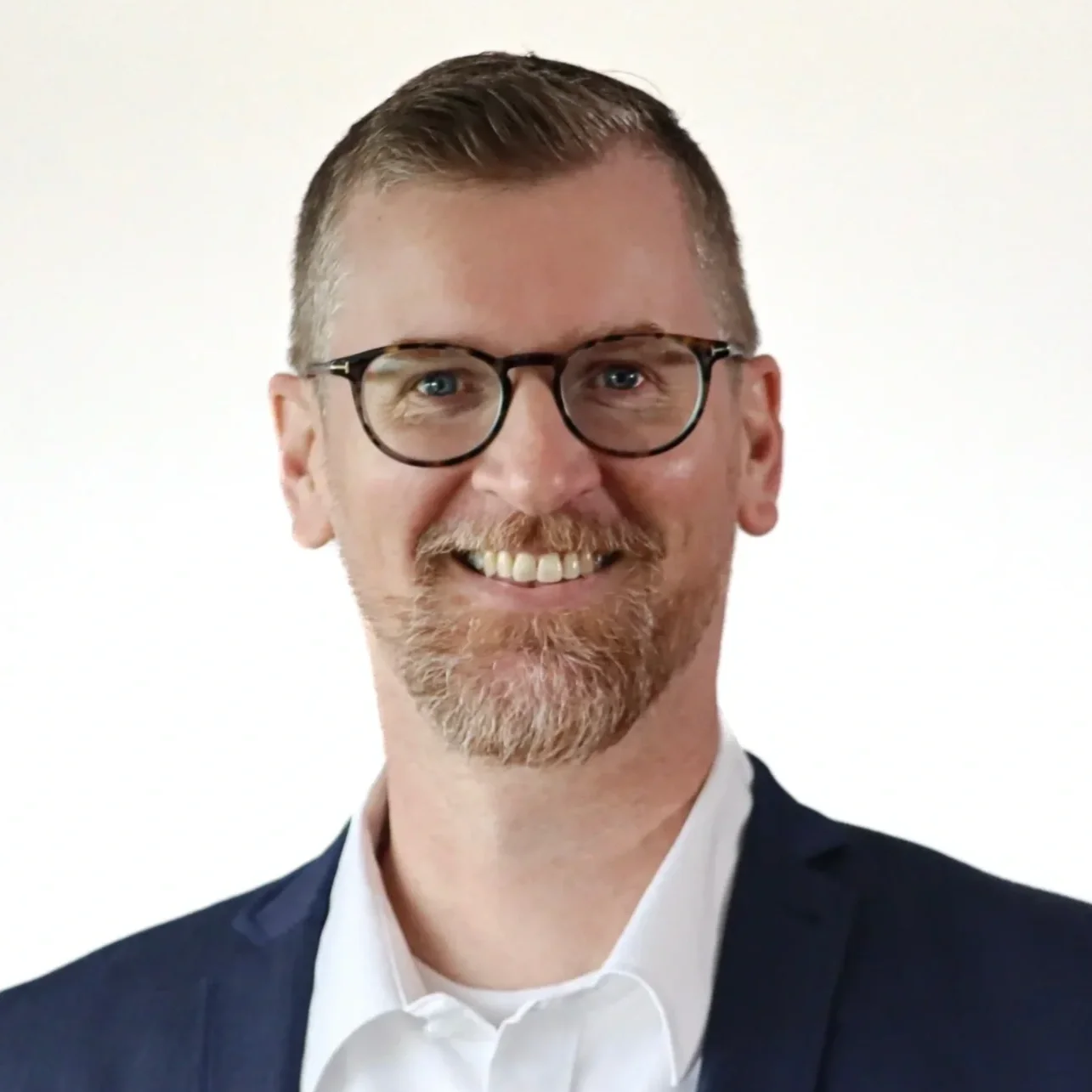 Headshot of a man with glasses, short brown hair, a beard, and mustache, smiling in formal attire with a white shirt and dark blazer.