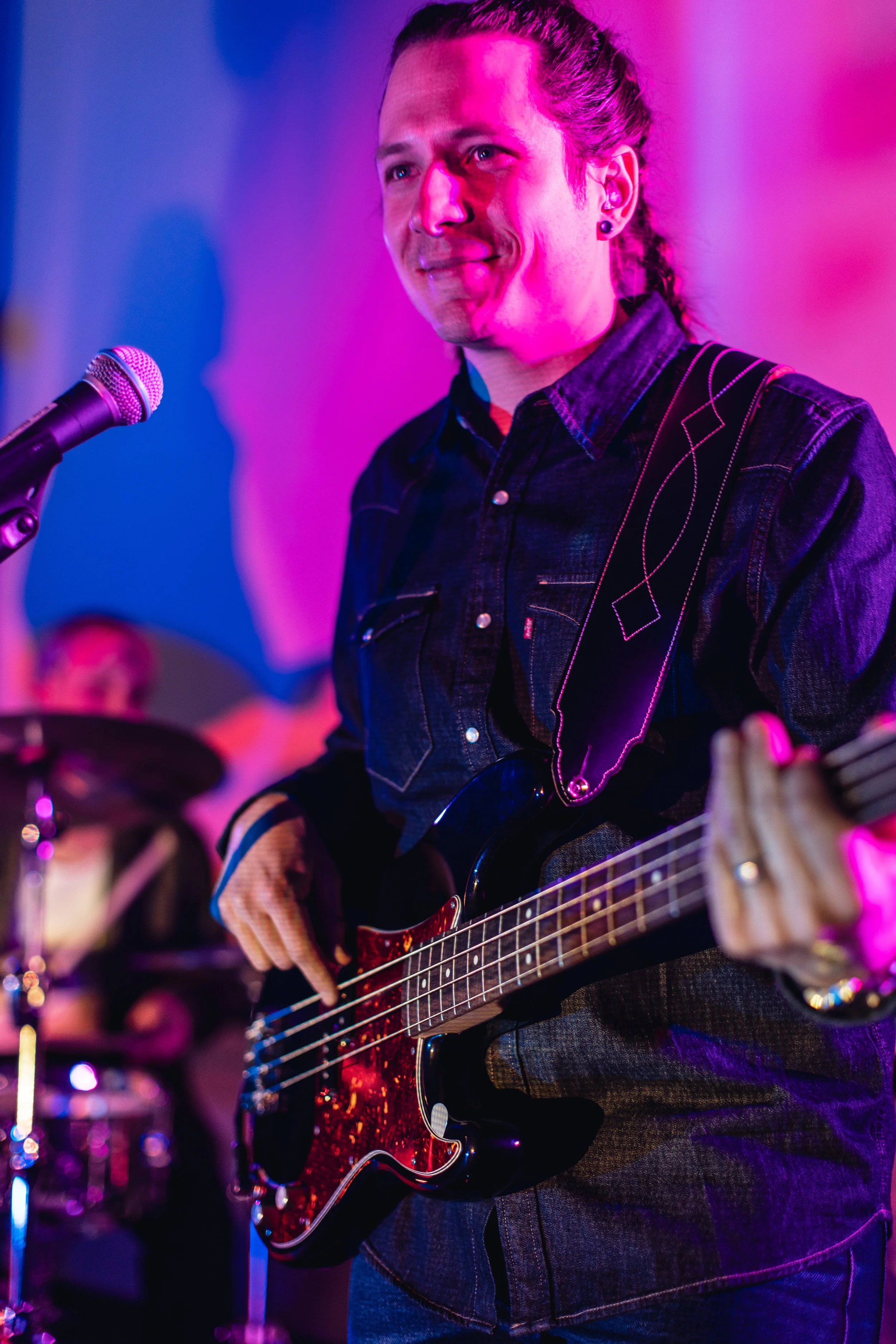 A person with long hair tied back, wearing earrings and a dark denim shirt, is playing a black electric guitar with a red and black body. They are standing near a microphone, with purple and pink stage lighting in the background.