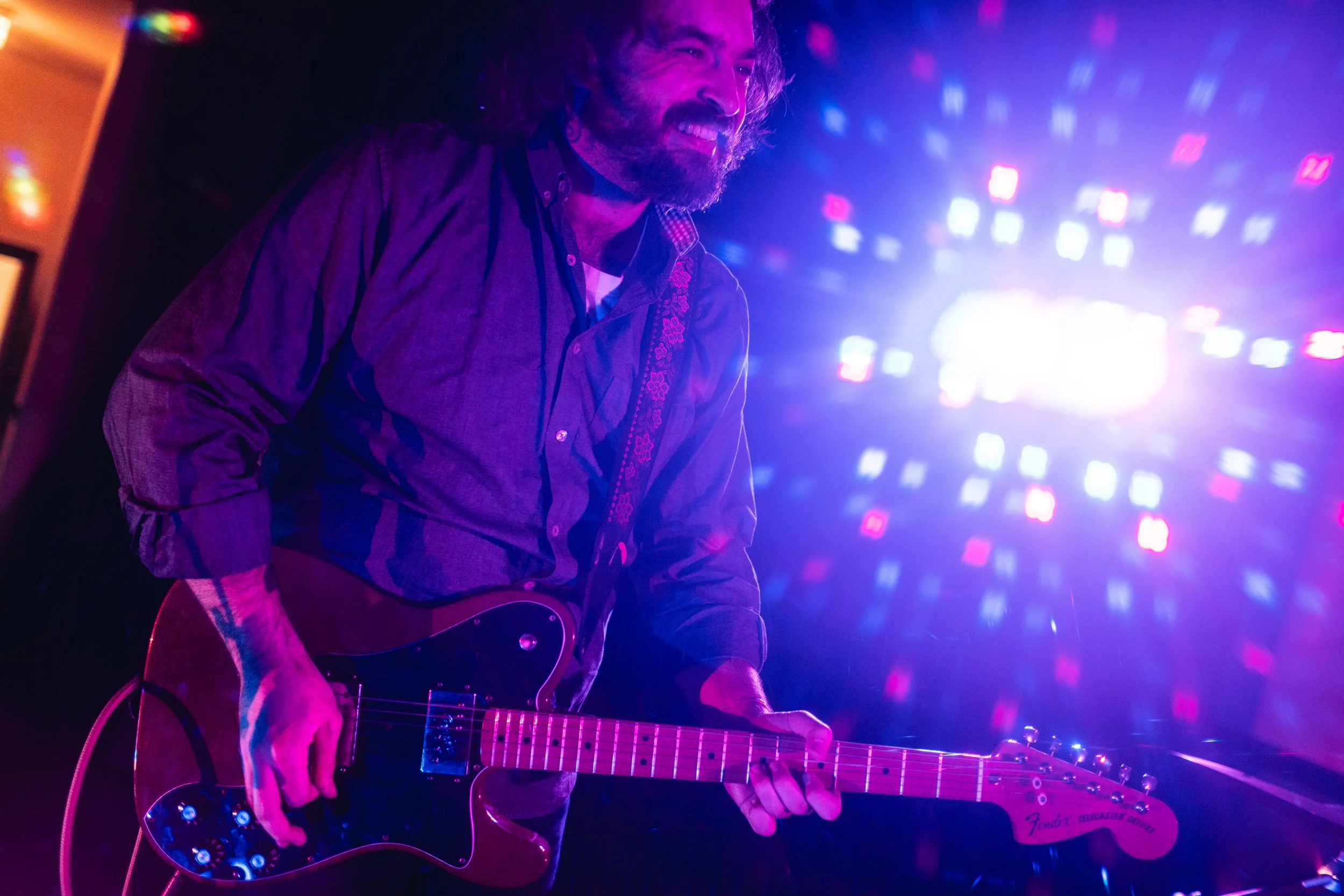 A man with a beard and long hair playing an acoustic guitar on stage, smiling, surrounded by colorful purple and blue lights.