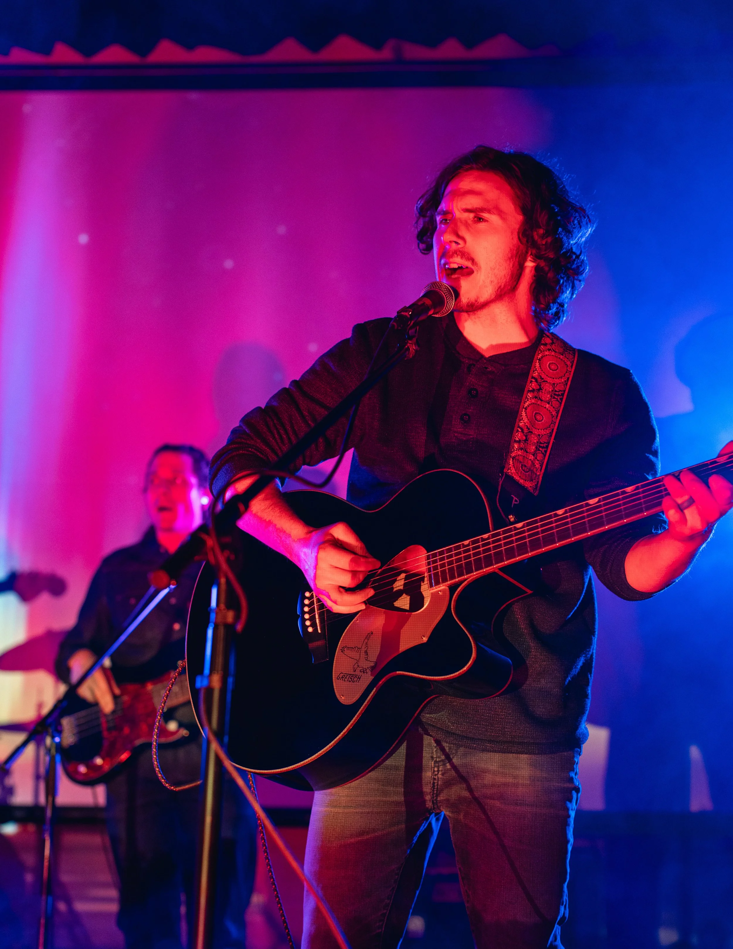 A man with curly dark hair and a beard singing into a microphone while playing an acoustic guitar on stage, with colorful stage lighting.