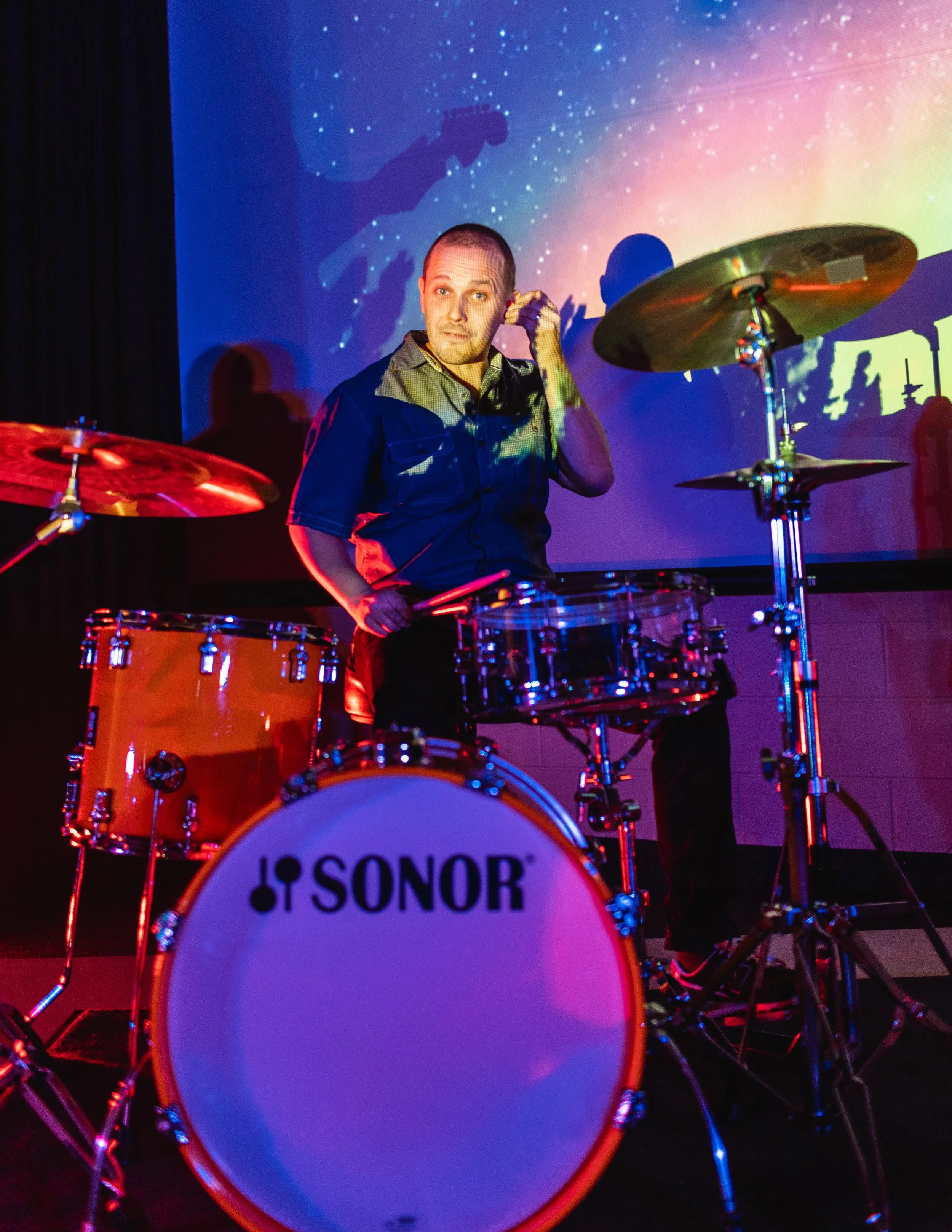 A person with a shaved head playing a drum set on stage with colorful lighting and a projection of a starry sky in the background.