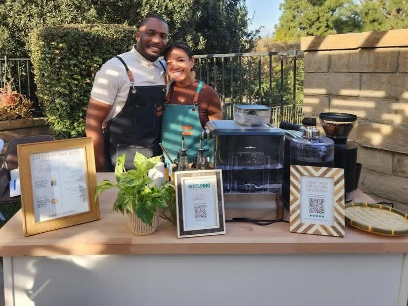Two people wearing aprons stand behind a coffee stand with machines, menu, plants, and QR code signs outside.