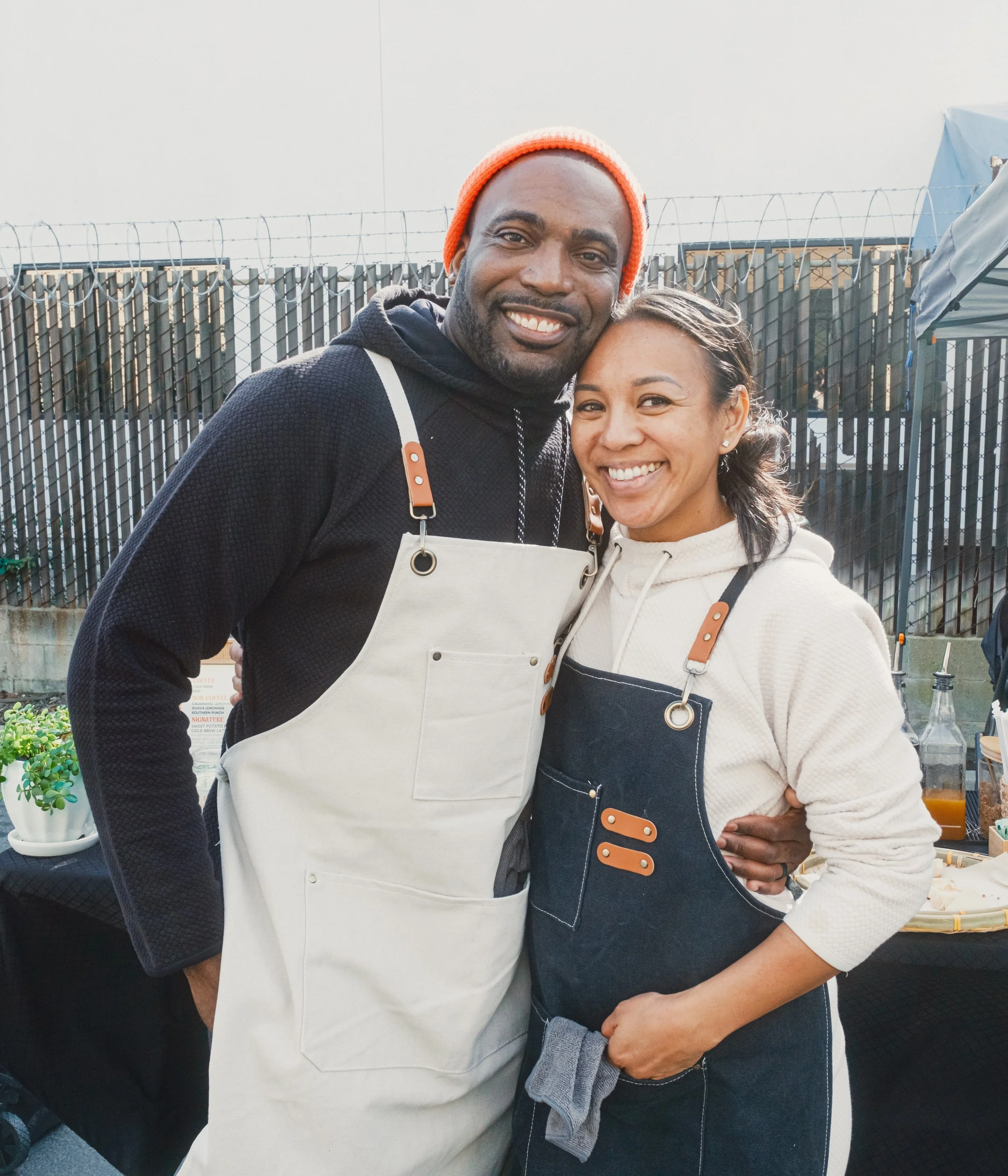 Two people wearing aprons, standing close together and smiling outdoors.
