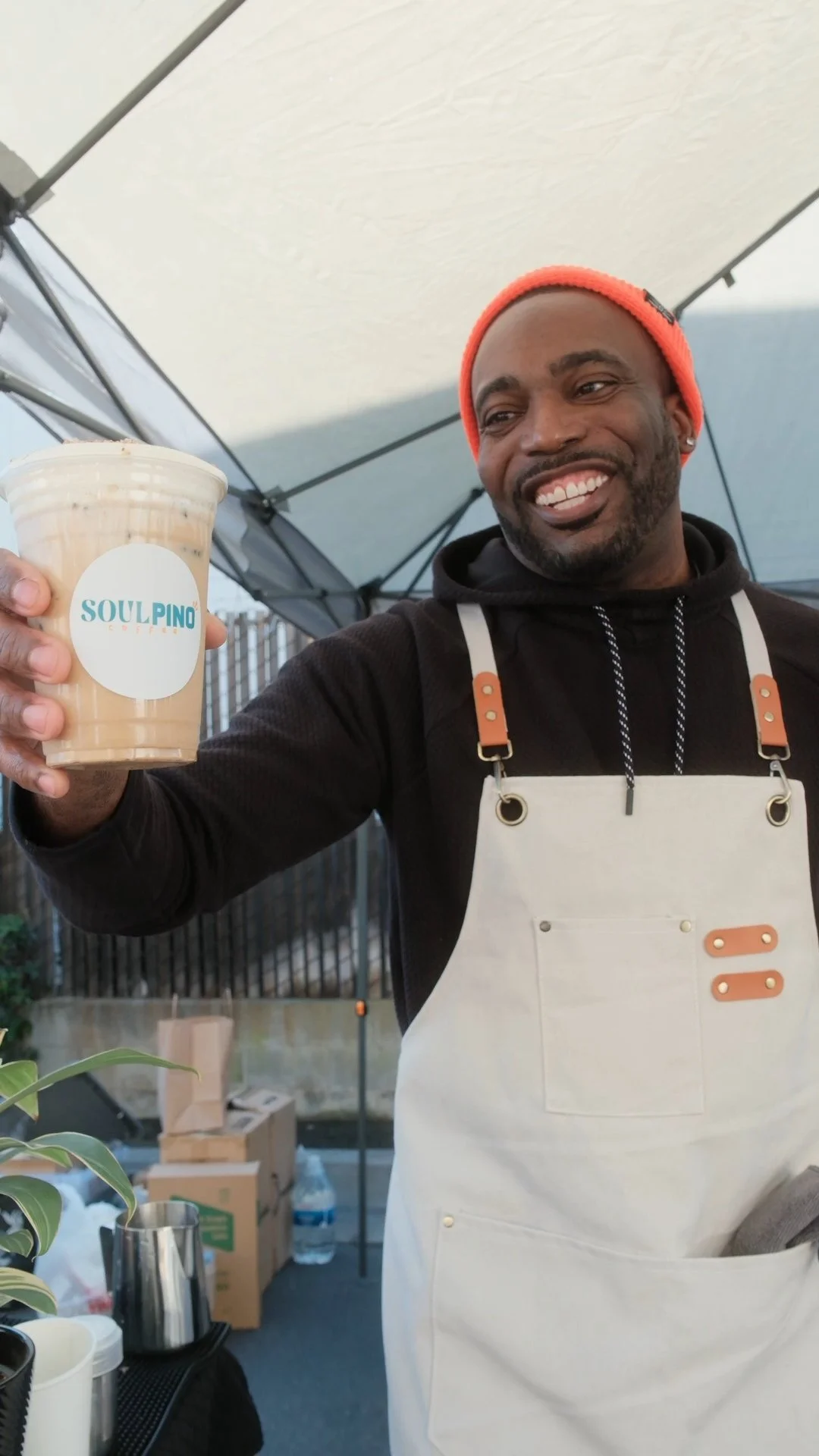 Man in a black hoodie and orange beanie holding a cup of iced coffee with "Soulpino Coffee" label, smiling in an outdoor market setting.