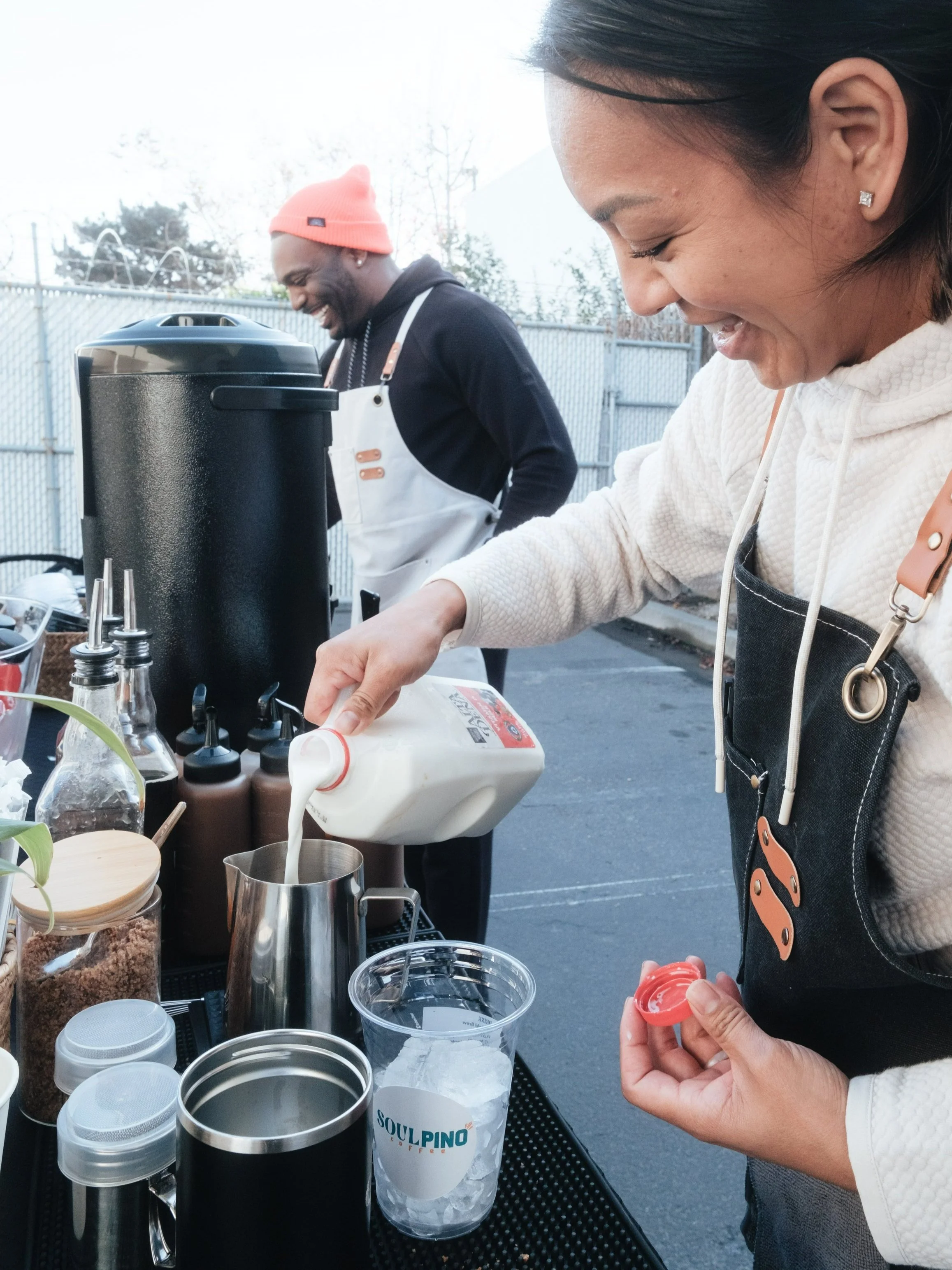 Two people preparing drinks outdoors; one is pouring milk into a metal pitcher, while various coffee-making supplies are arranged on a table.