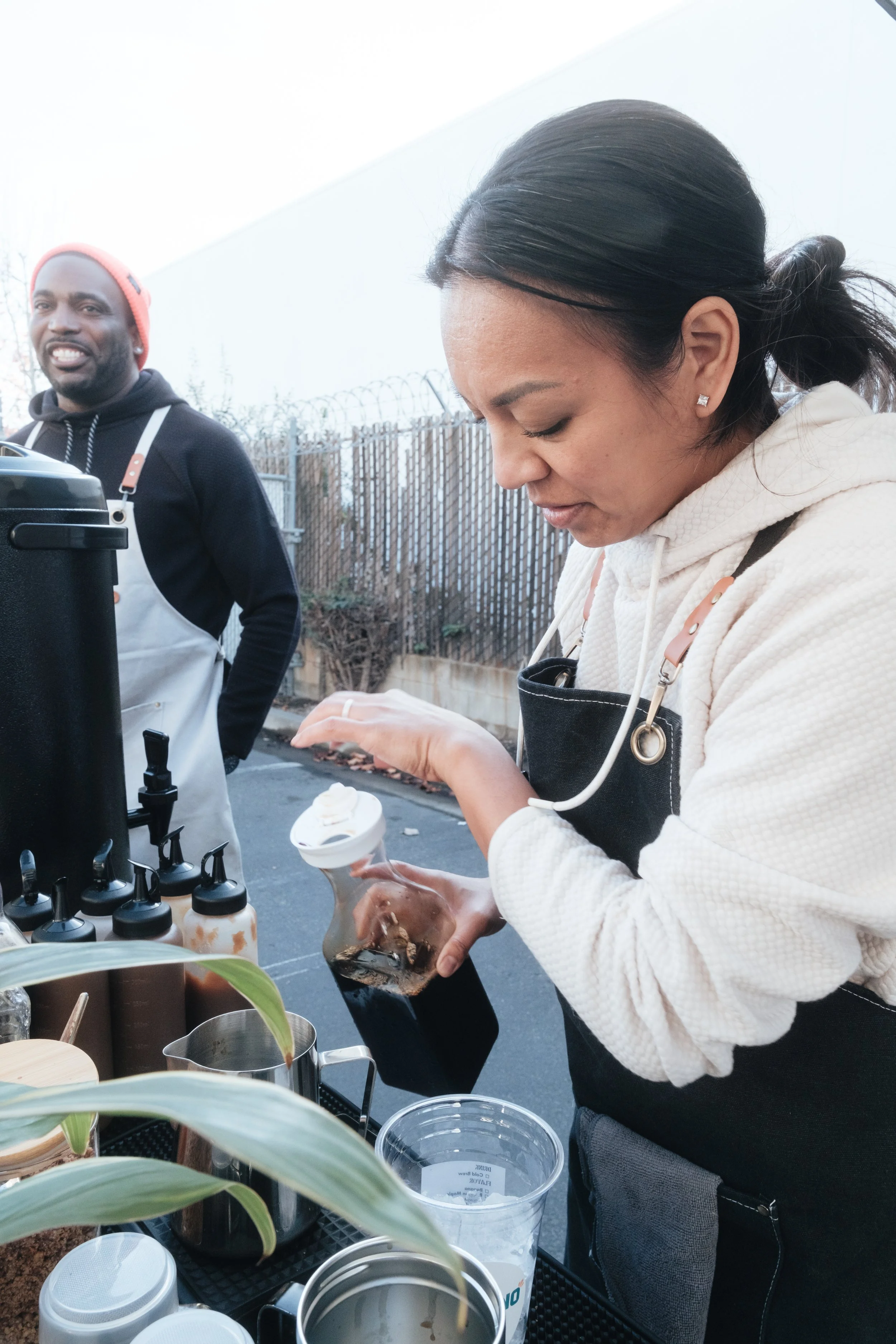 Two people making coffee outdoors, one pouring liquid from a bottle, with various coffee supplies on a table, including syrup dispensers and a metal pitcher.