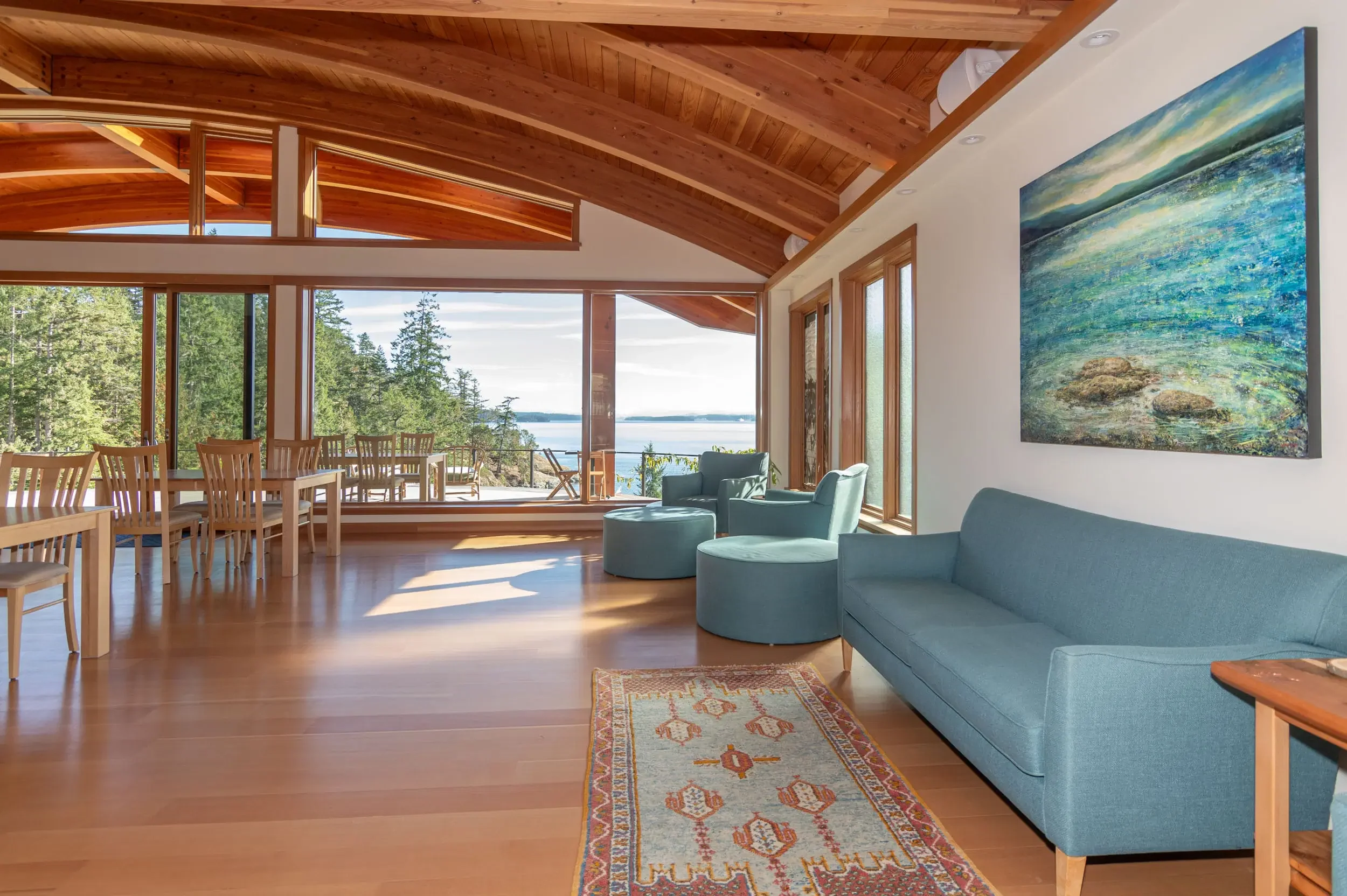 Living room with large windows overlooking a forest and water, wooden ceiling, blue sofa, matching armchairs, and colorful area rug.