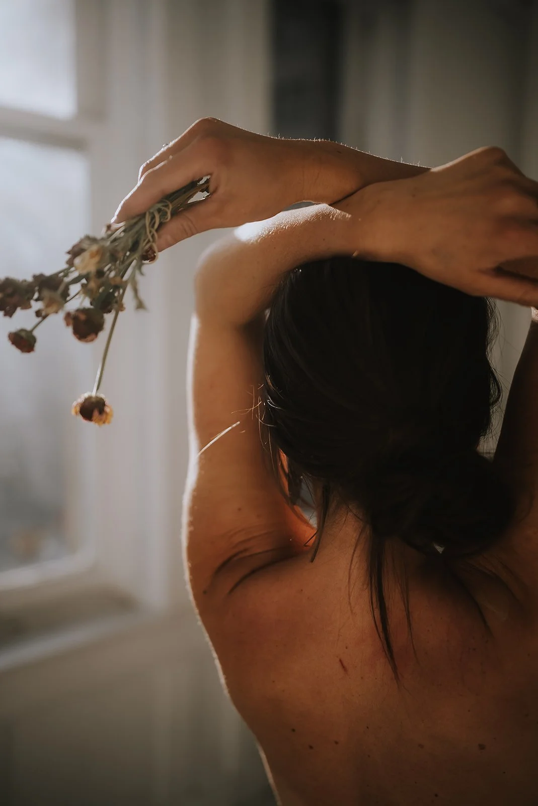 Close-up of a woman with dark hair holding dried flowers behind her head, with sunlight coming through a window.