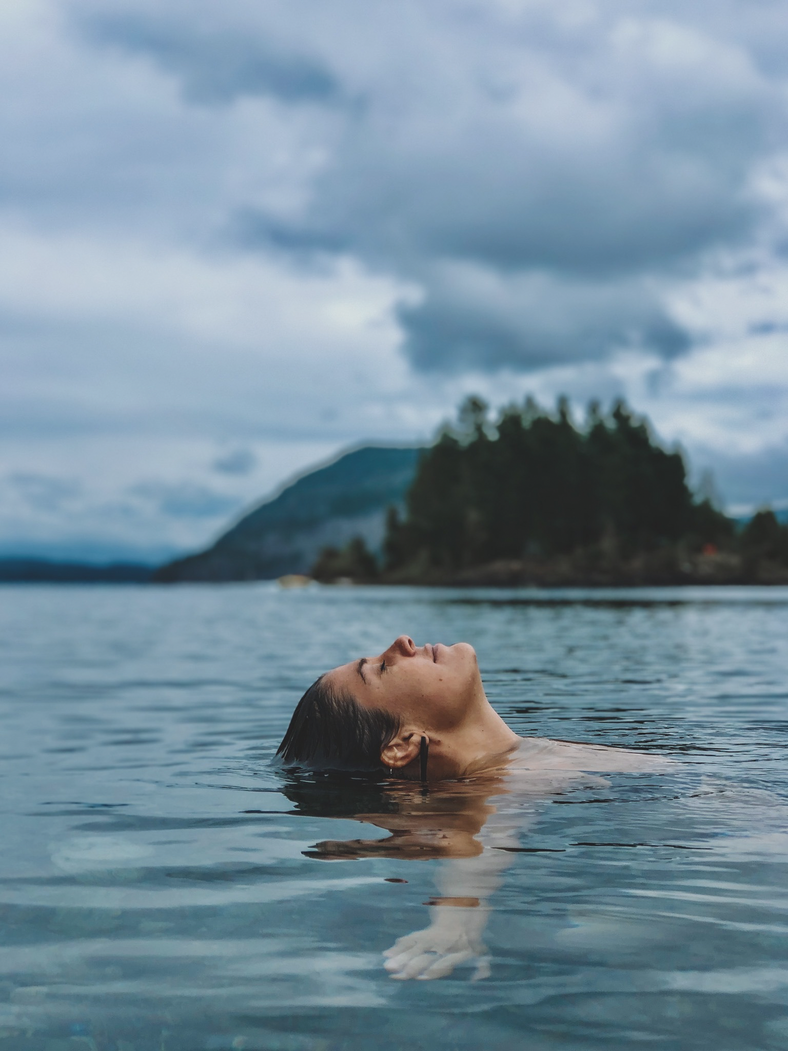 A woman swimming in a lake with a mountainous landscape and cloudy sky in the background.