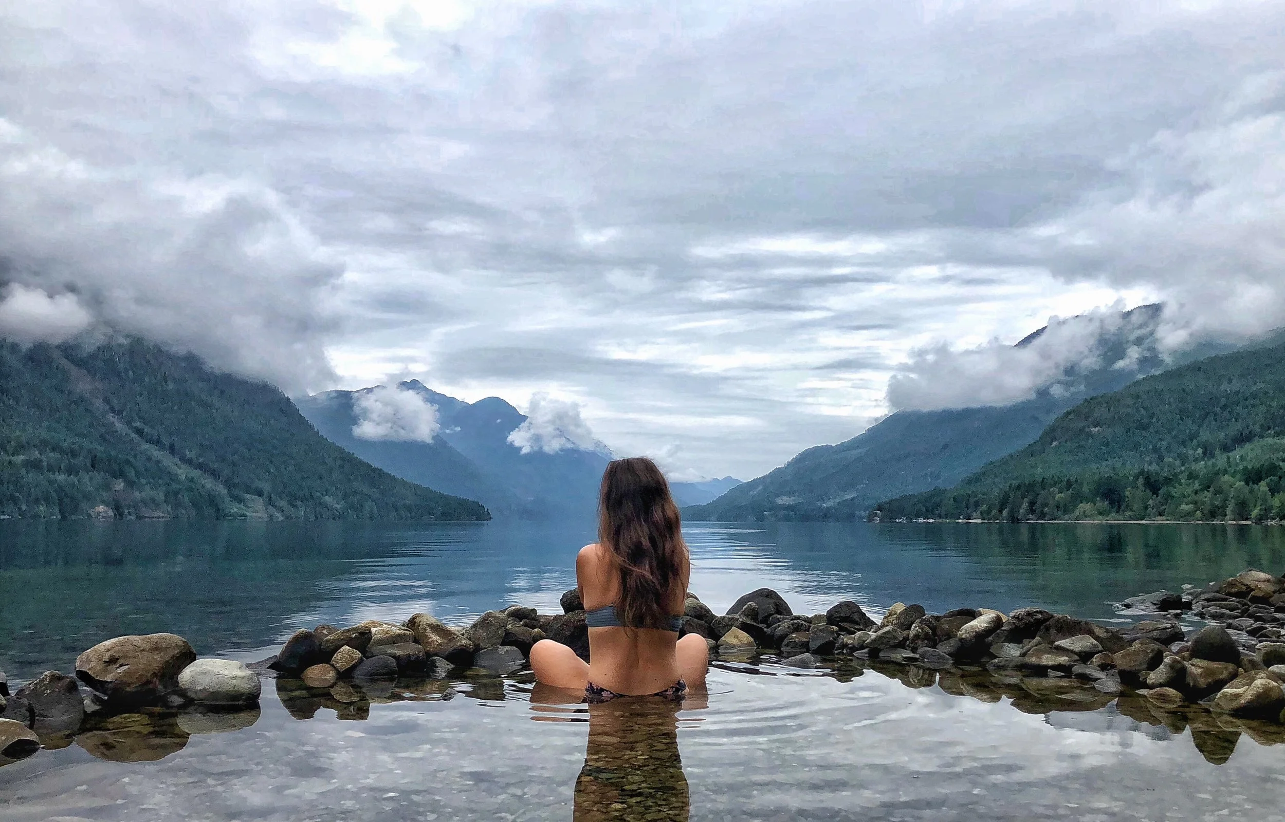 A woman with long brown hair sitting in a natural hot spring or pool by a mountain lake, surrounded by rocky edges, with a view of mountains, water, and cloudy sky in the background.