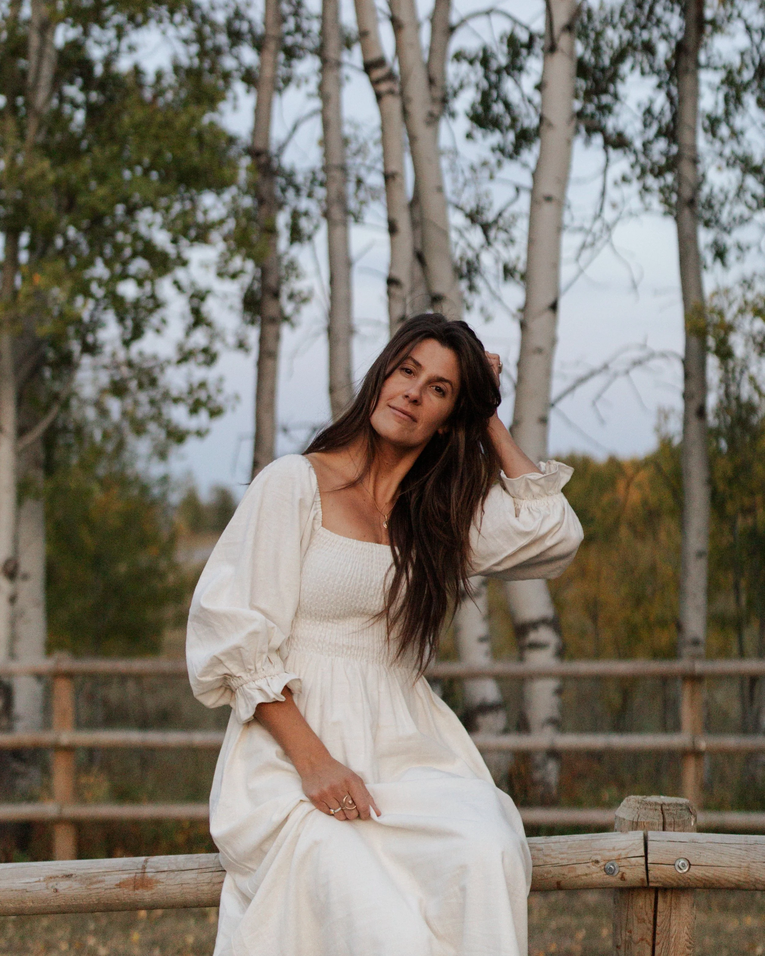 A woman in a white dress sitting on a wooden railing outdoors with trees in the background.