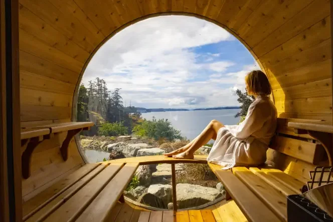 A person sitting inside a wooden, circular structure with an open front, overlooking a scenic body of water, rocks, trees, and a cloudy sky.