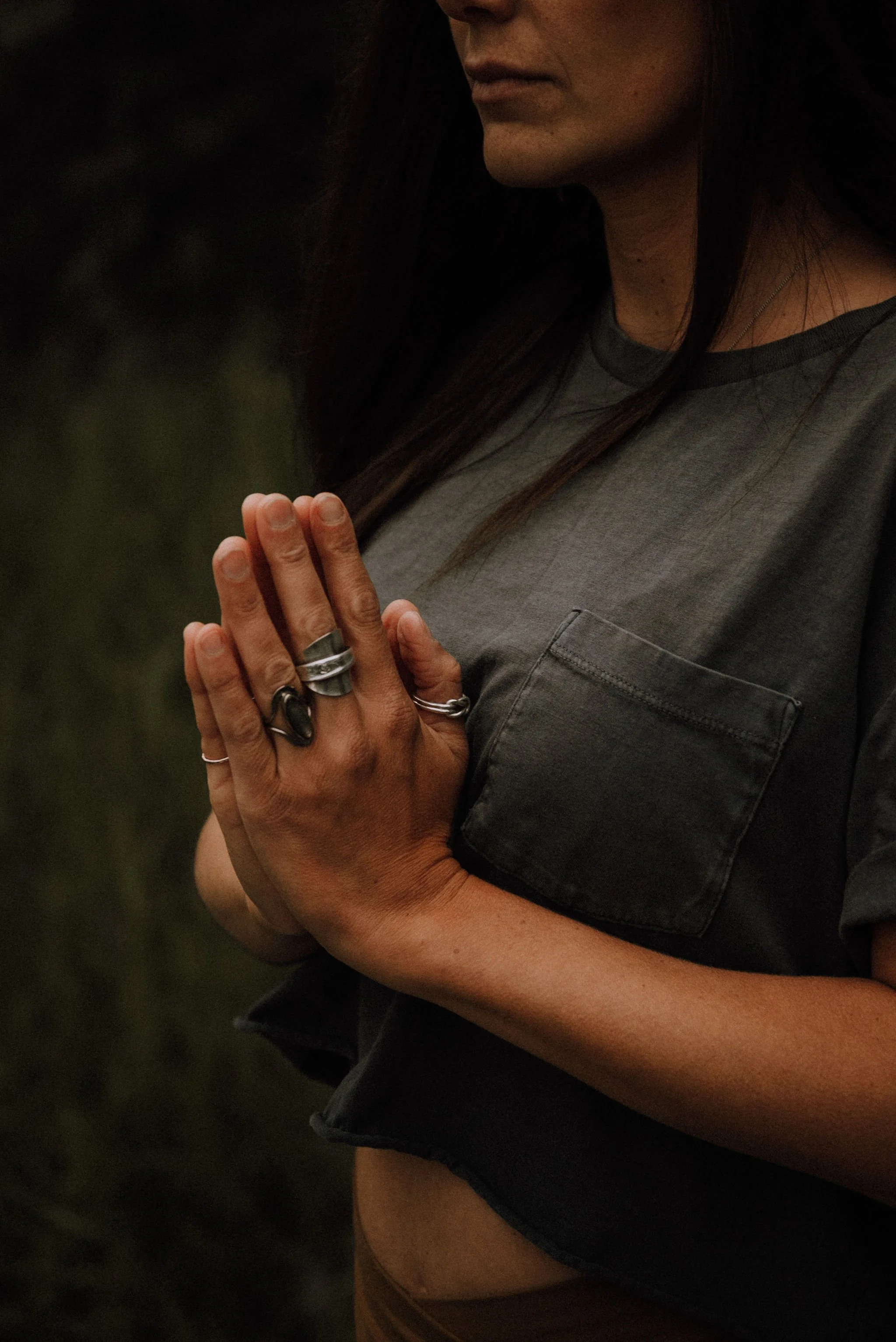 A woman with long dark hair, wearing a dark grey T-shirt with a chest pocket, has her hands pressed together in a prayer or meditation pose.