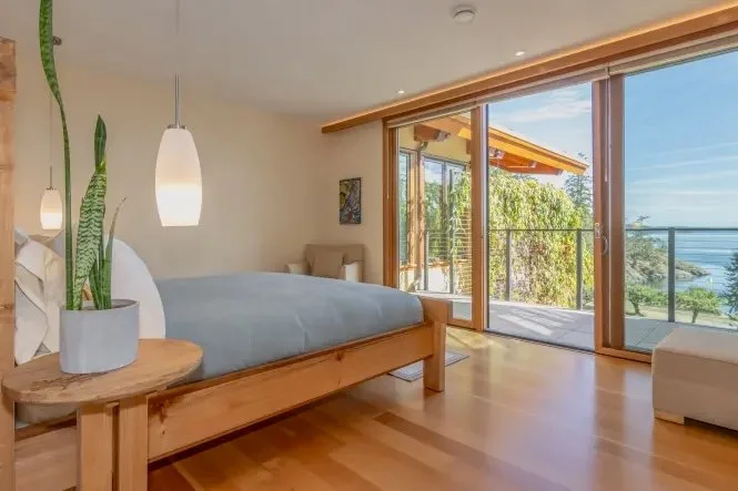 A bedroom with a wooden bed, potted plant on a side table, hanging pendant lights, large sliding glass doors leading to a balcony, ocean view, and trees outside.