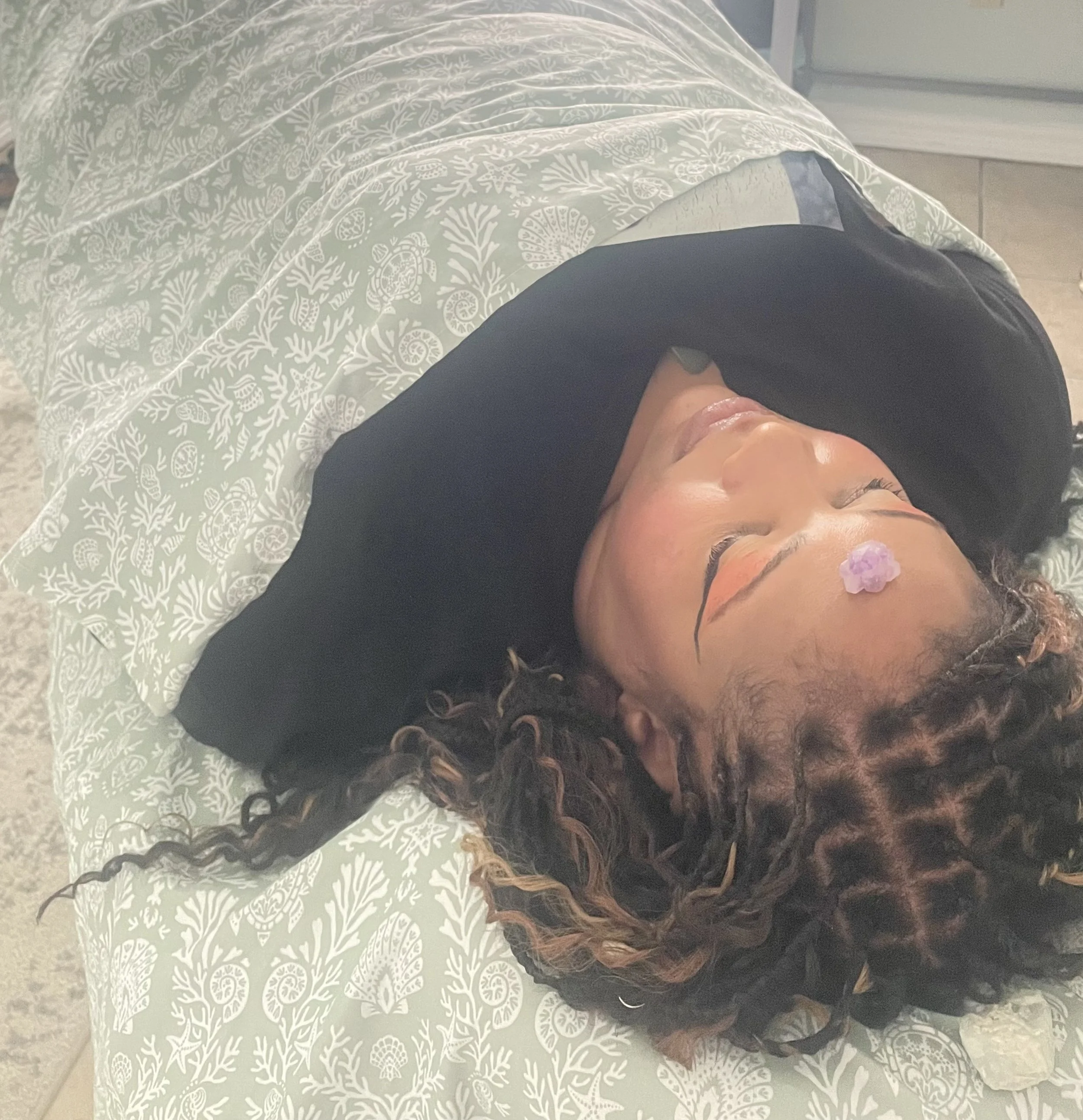 A woman with curly hair lying on her back on a bed with a patterned bedspread, with a small purple Amethyst crystal on her forehead, resting peacefully with her eyes closed.