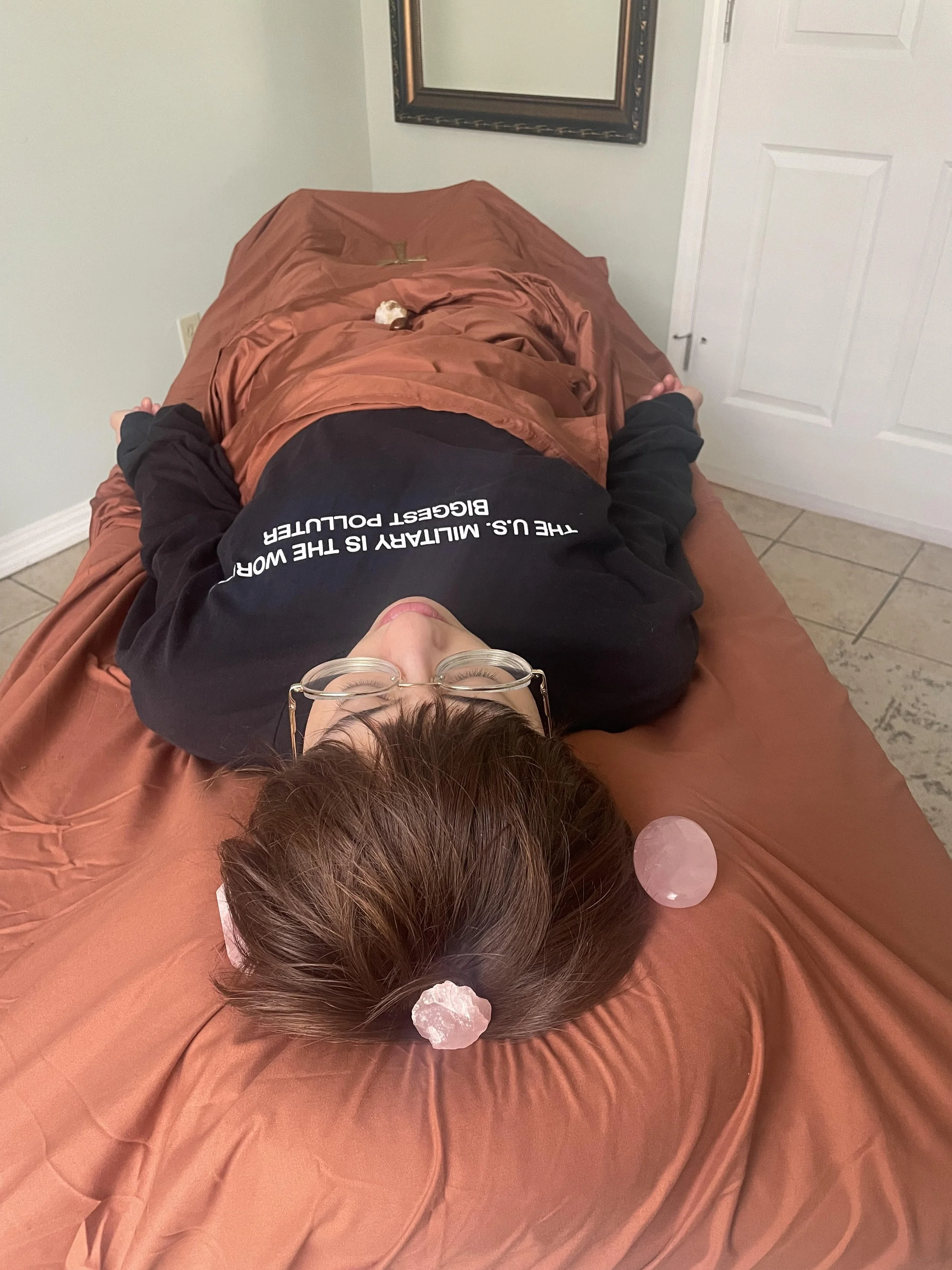 Person lying face up on a massage table with rose quartz stones on their head and a black shirt that says 'The U.S. Military is the worst biggest polluter' in a room with white walls, a mirror, and a white door.