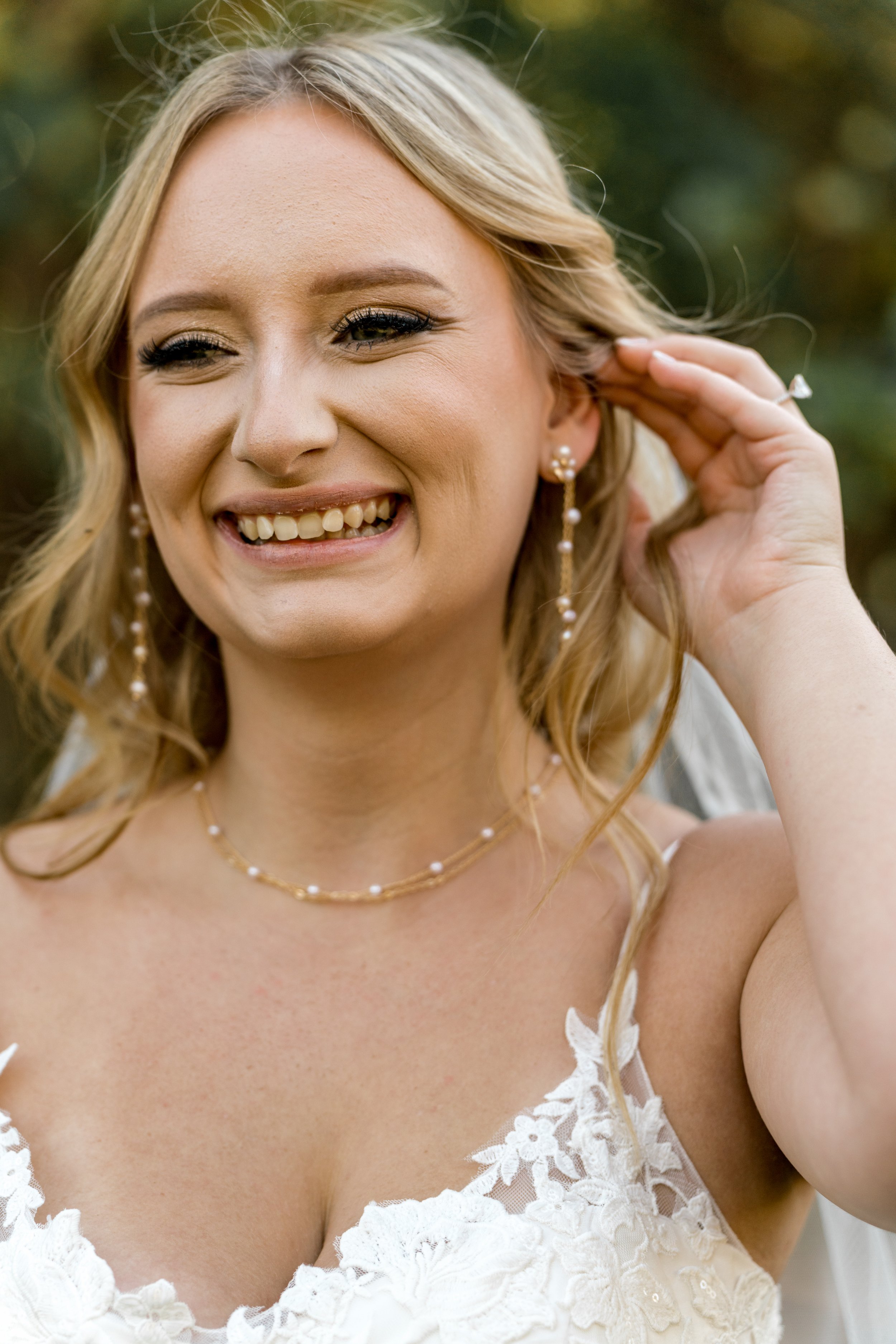Smiling bride with blonde hair, wearing a white lace wedding dress and pearl jewelry, adjusting earring outdoors.