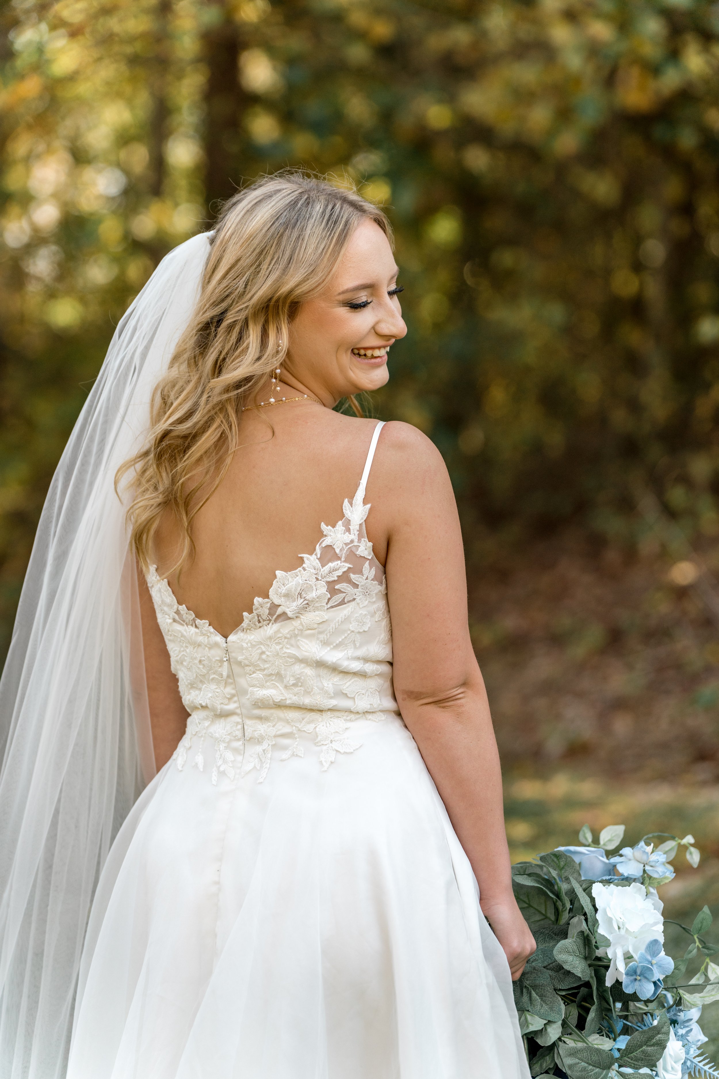 Bride with long veil and lace wedding dress holding blue flowers, outdoors.