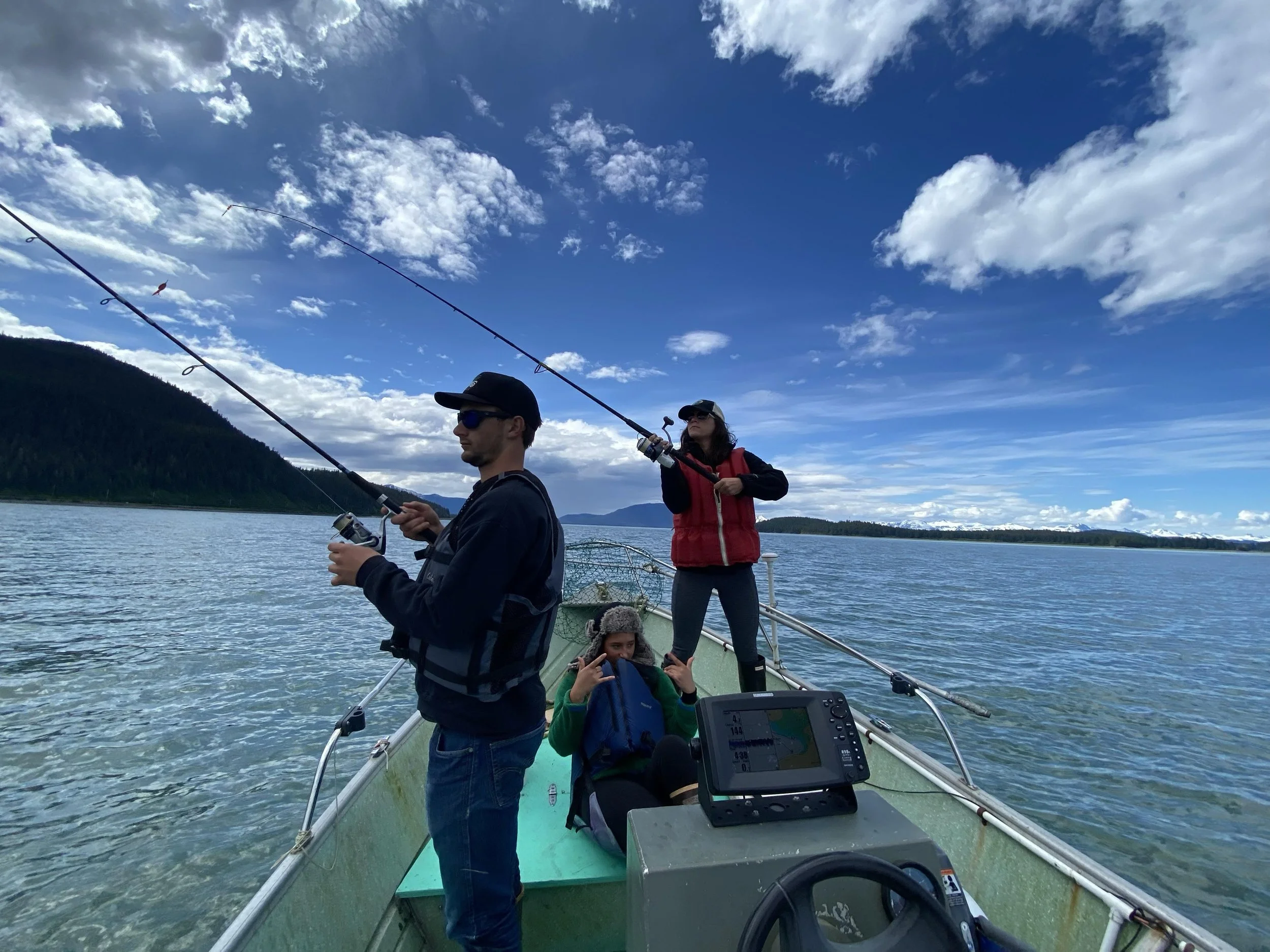 Three people fishing on a boat on a lake during daytime, with mountains and cloudy sky in the background.