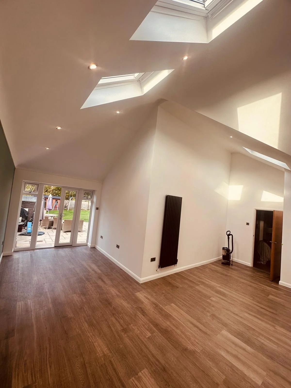 Empty living room with wooden flooring, white walls, skylights on the sloped ceiling, and large glass doors leading to an outdoor patio with garden furniture.