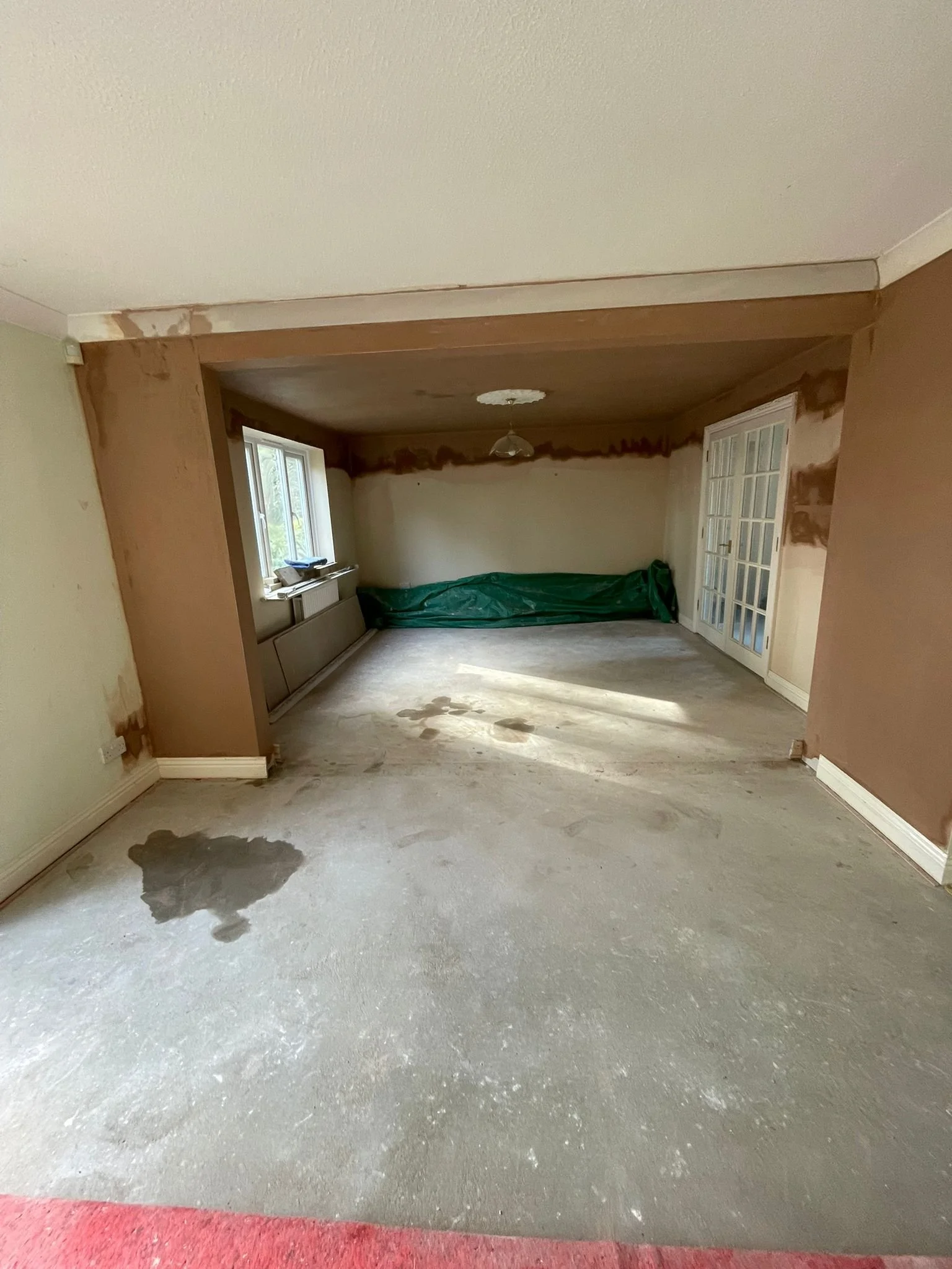 Room undergoing renovation with unfinished floor, partially painted walls, and a window on the left, door on the right, with some construction materials and a green tarp.