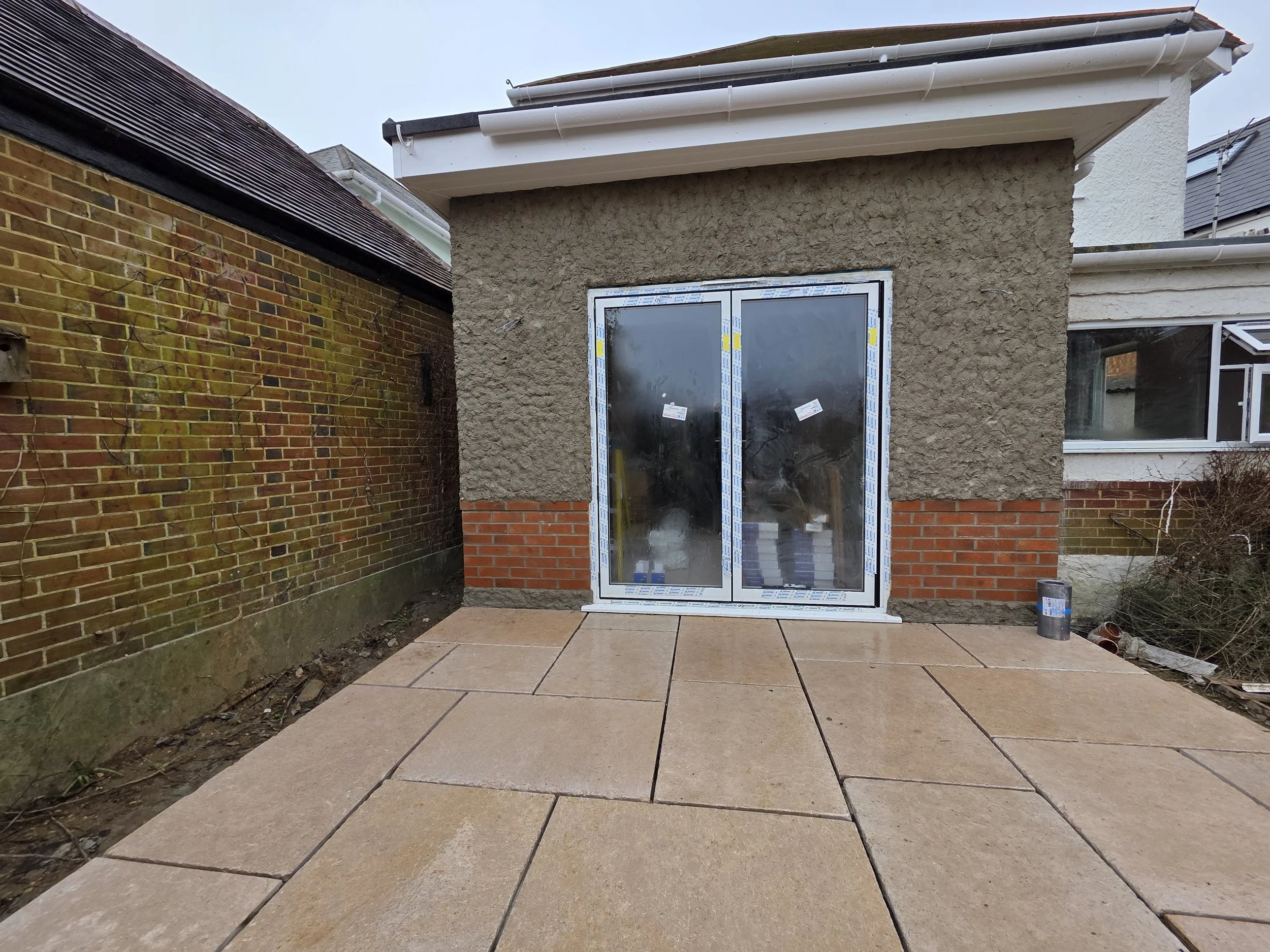 A house with a small extension under construction showing a glass door and a window, surrounded by brick and textured wall, with paving stones on the ground and a brick wall on the left side.