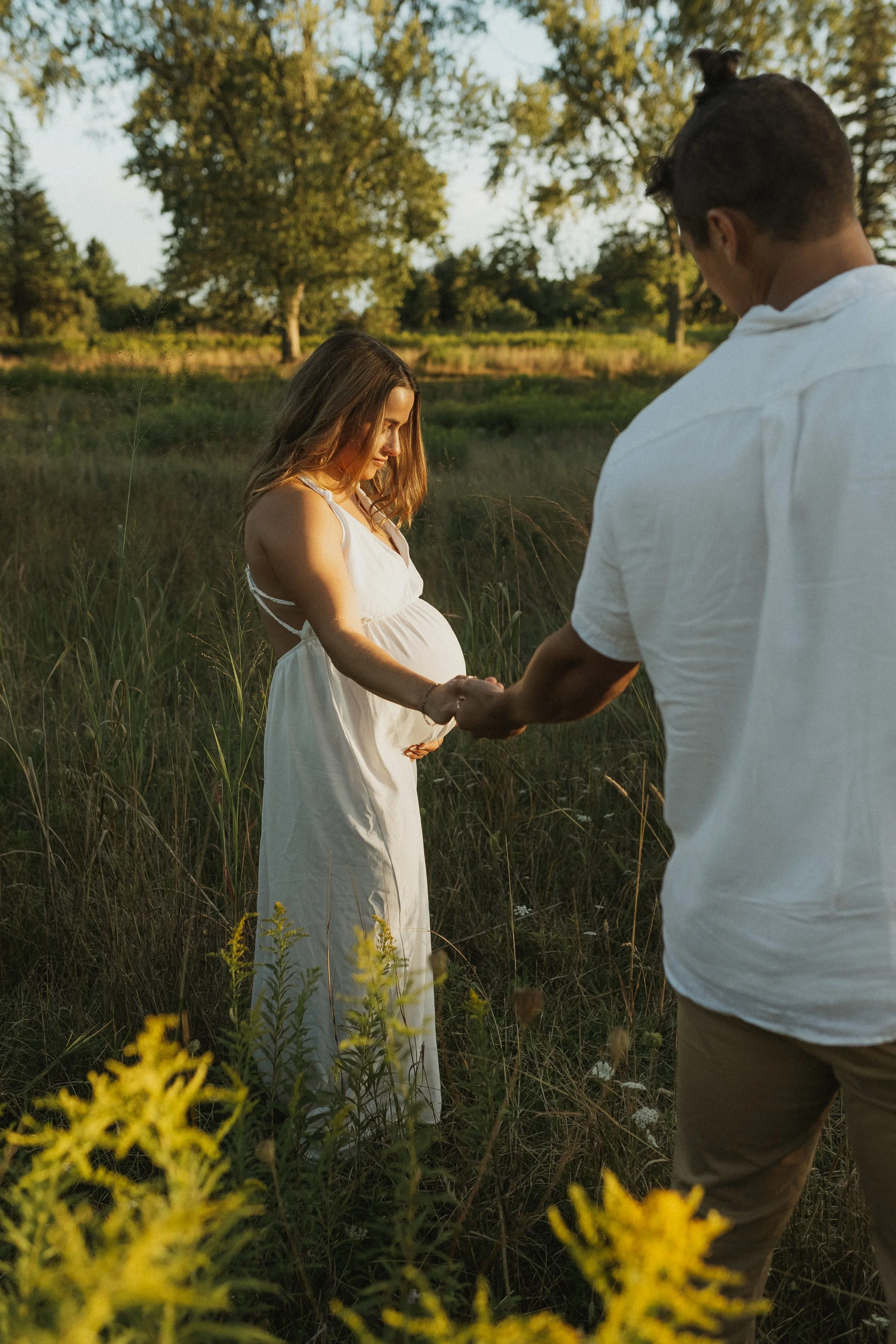 A pregnant woman in a white dress holding hands with a man in a casual white shirt in a grassy field during sunset.