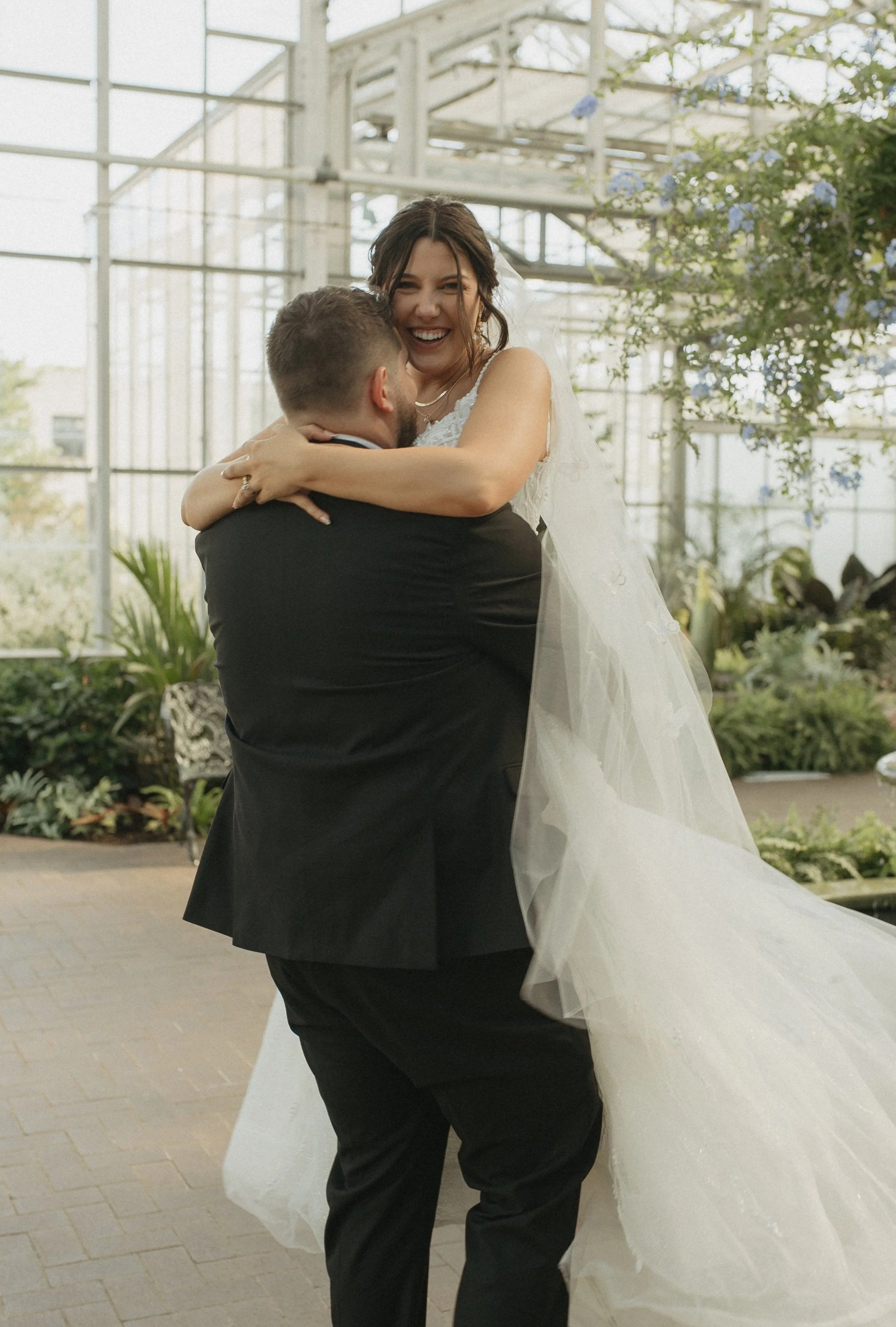 A bride and groom dancing inside a greenhouse with glass walls and lush plants, with the groom lifting the bride, who is smiling.