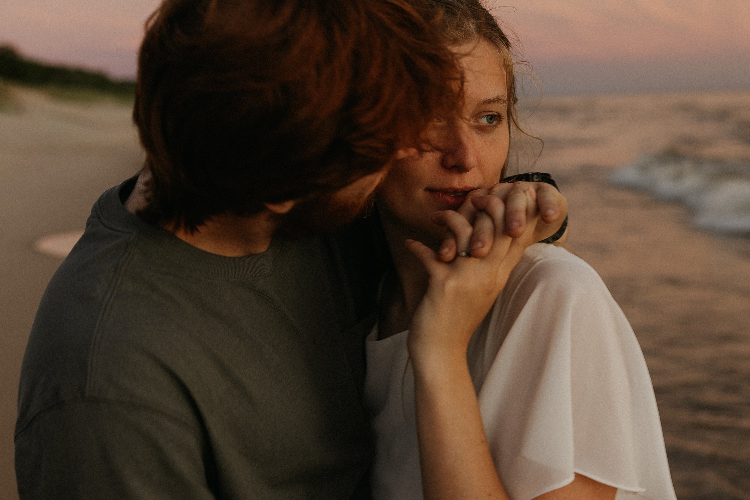 A romantic scene of a man and woman on the beach at sunset, close together, fingers interlocked, as the man kisses the woman on the cheek.