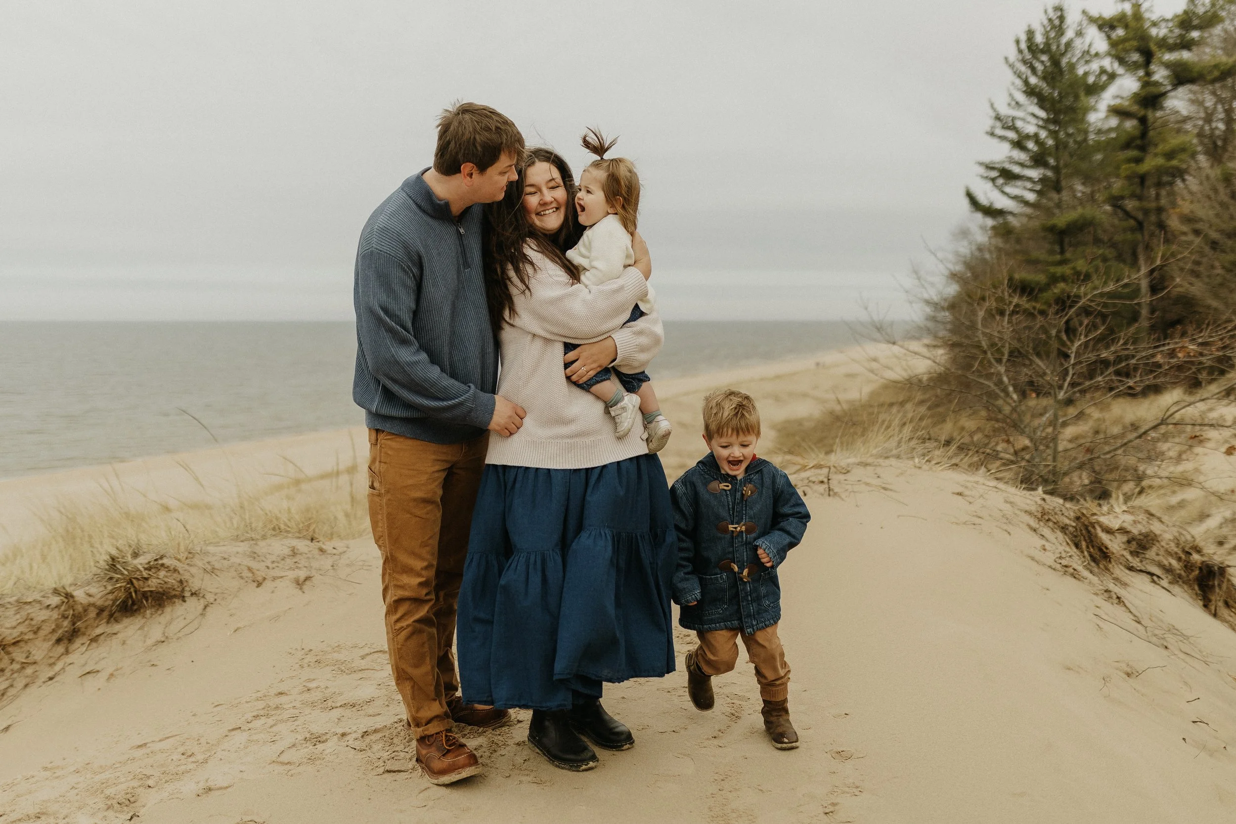 A family of four walking on a sandy beach with grass and trees, enjoying a cloudy day near the water.