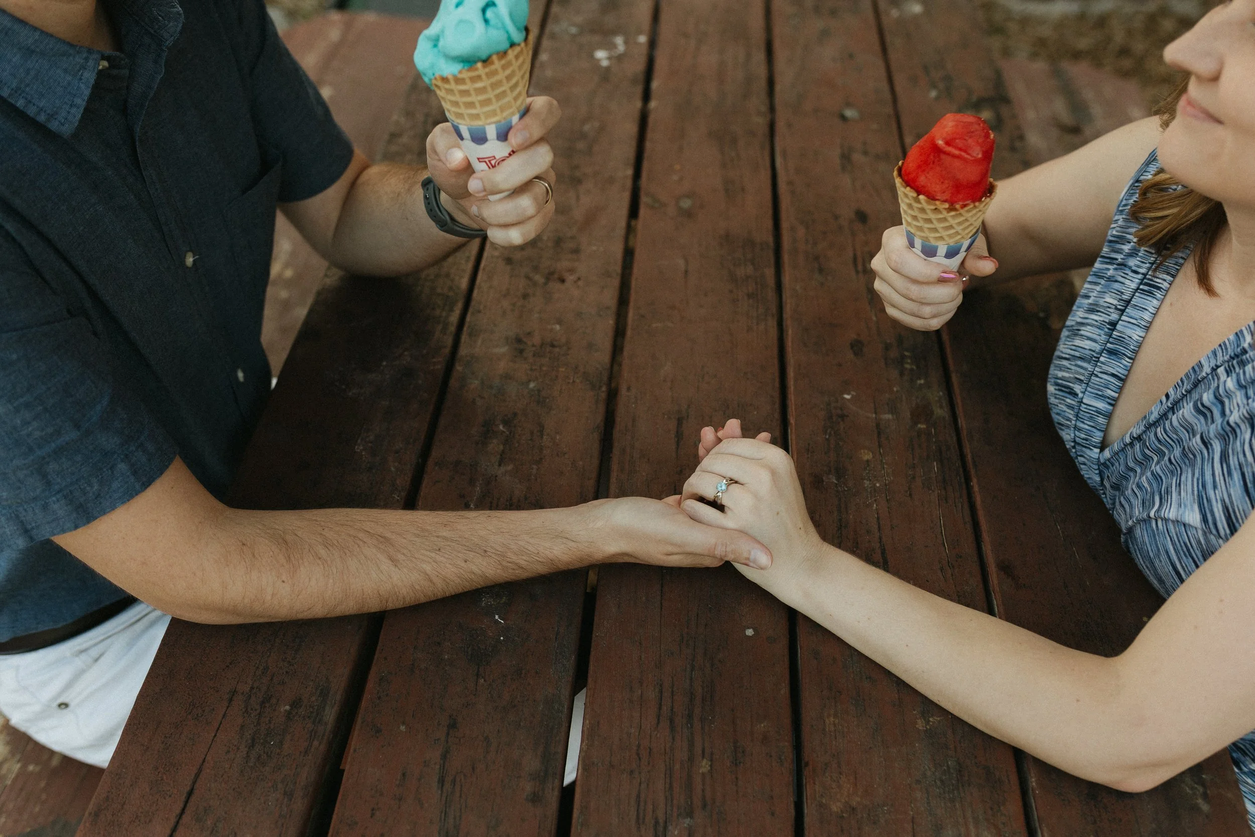 A man and woman sitting at a wooden picnic table holding ice cream cones, with their hands clasped.