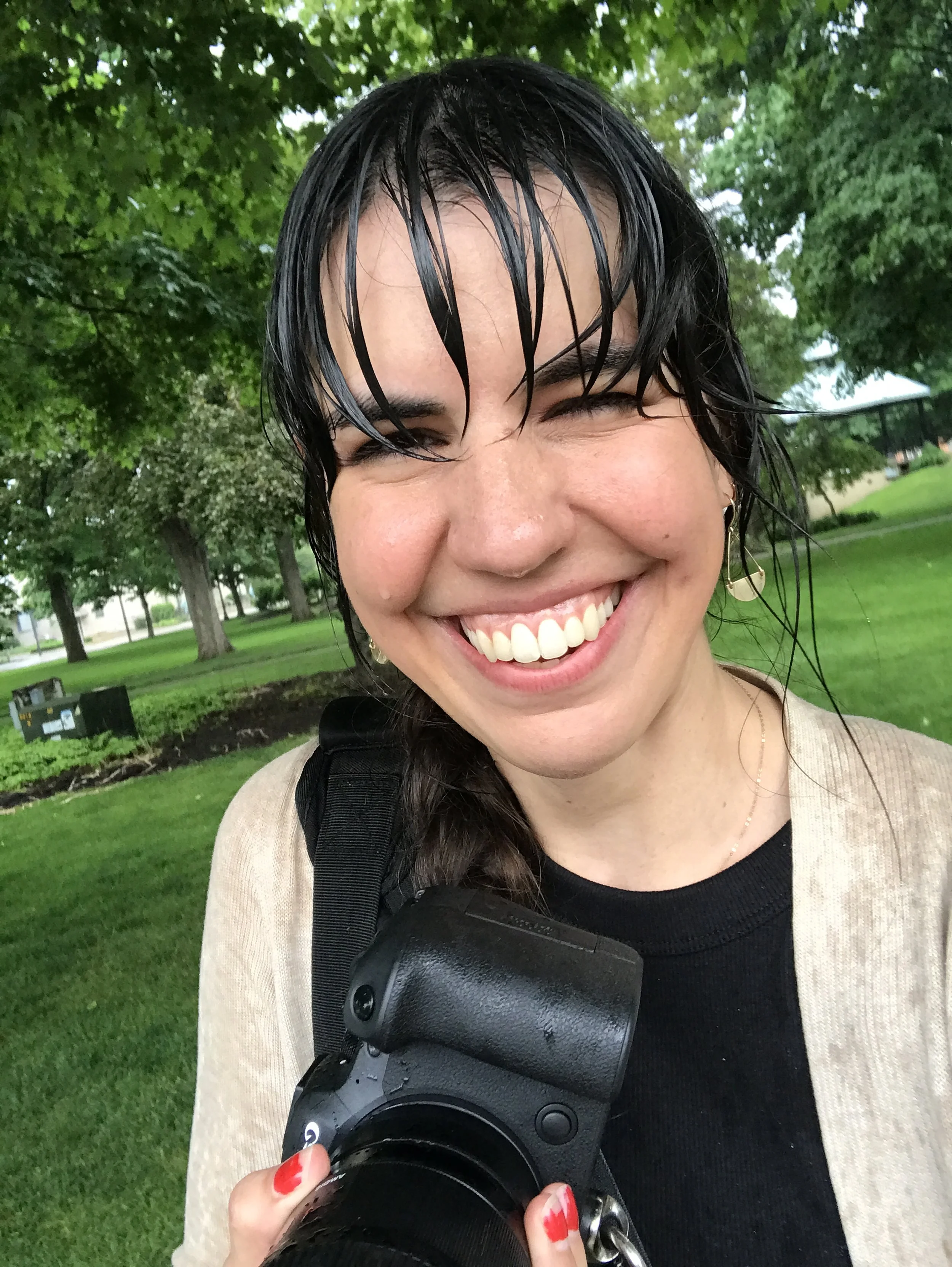 Smiling woman with wet black hair holding a camera outdoors in a park with green trees and grass.