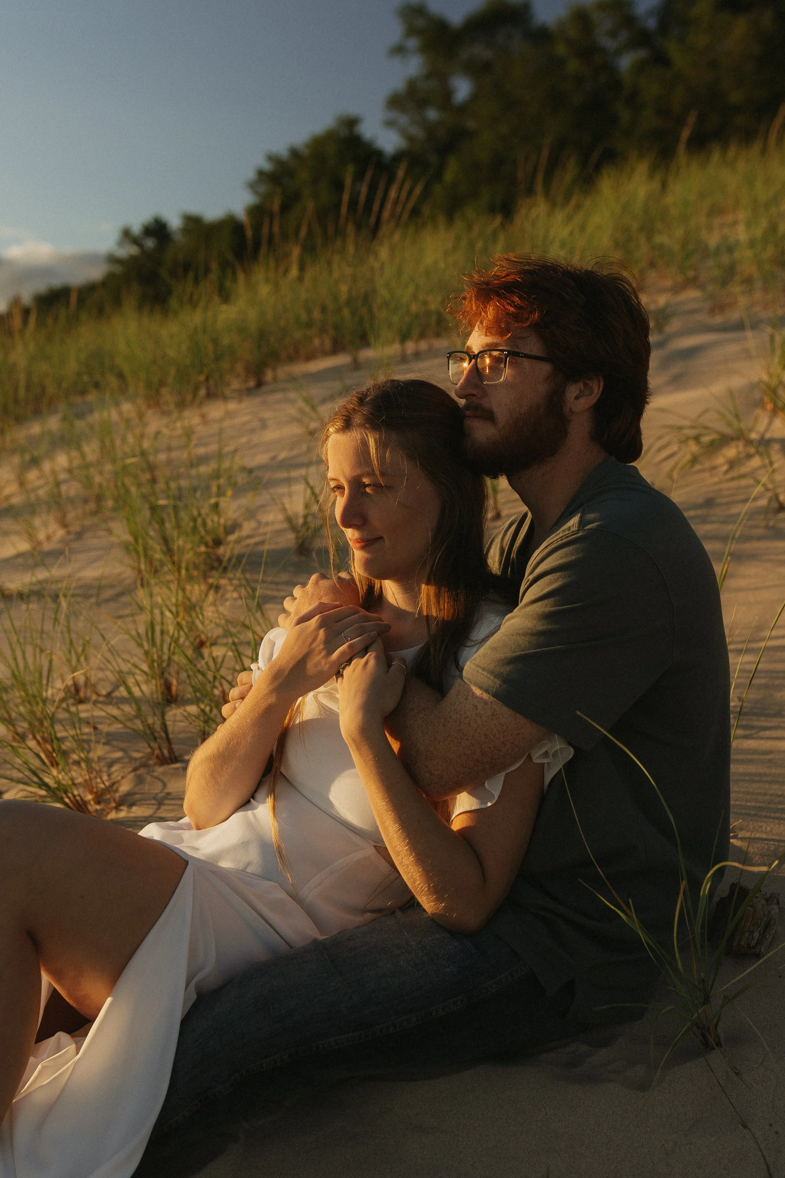 A man and woman sitting on a sandy beach, embracing at sunset, with dune grass and trees in the background.