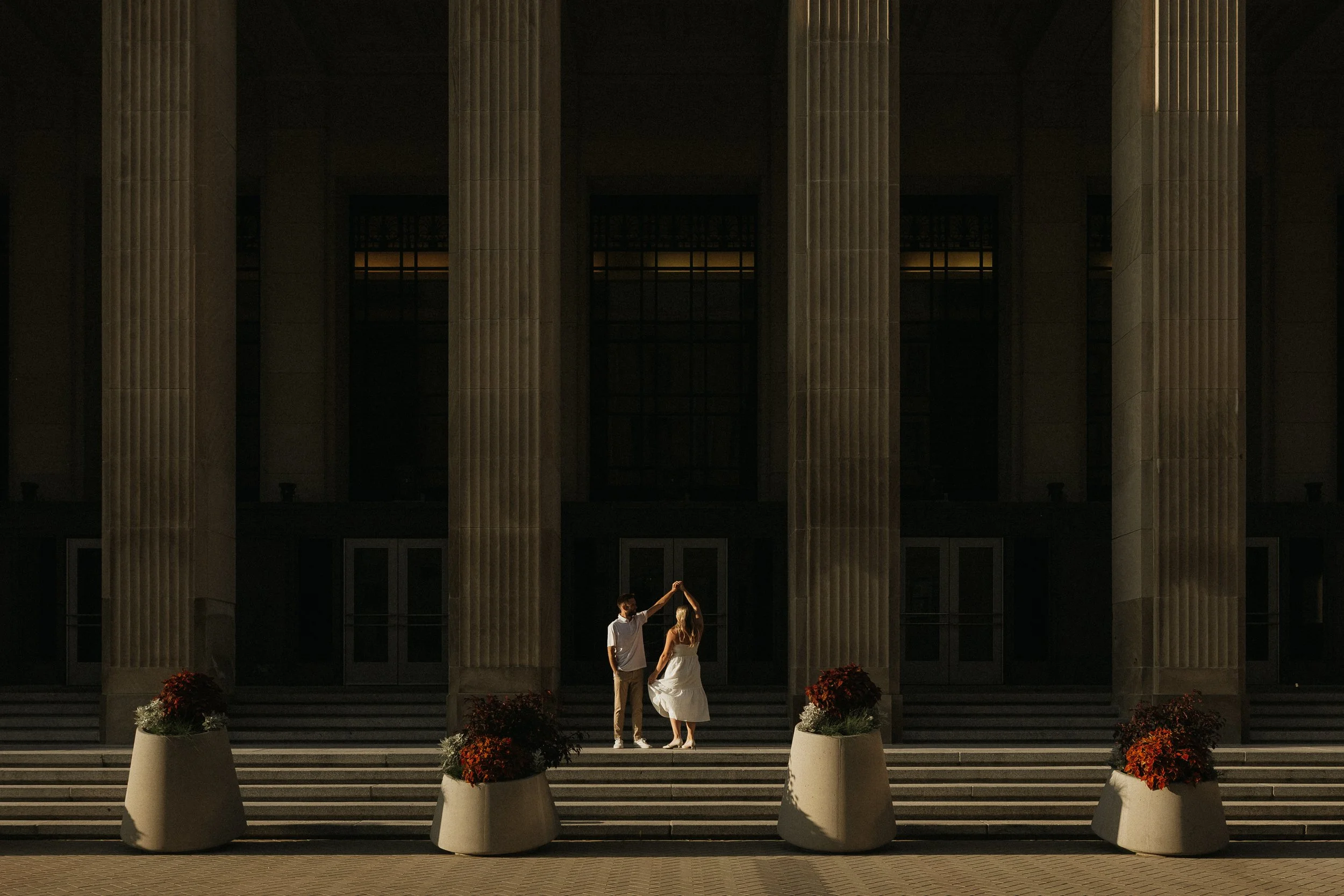 Two people dancing on front steps of a large building at sunset with tall columns and planters with flowers.