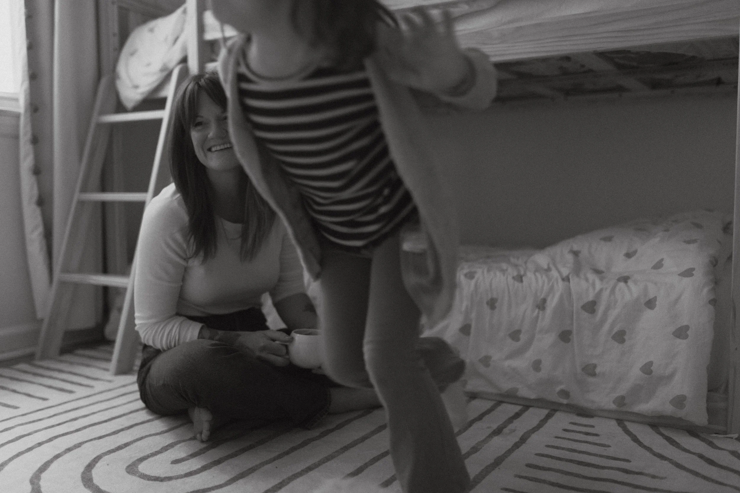 Two people, one sitting on the floor and the other partially visible, in a room with bunk beds. The person sitting has long dark hair, is smiling, and holding a cup. The room has a patterned rug and a bed with heart-patterned bedding.