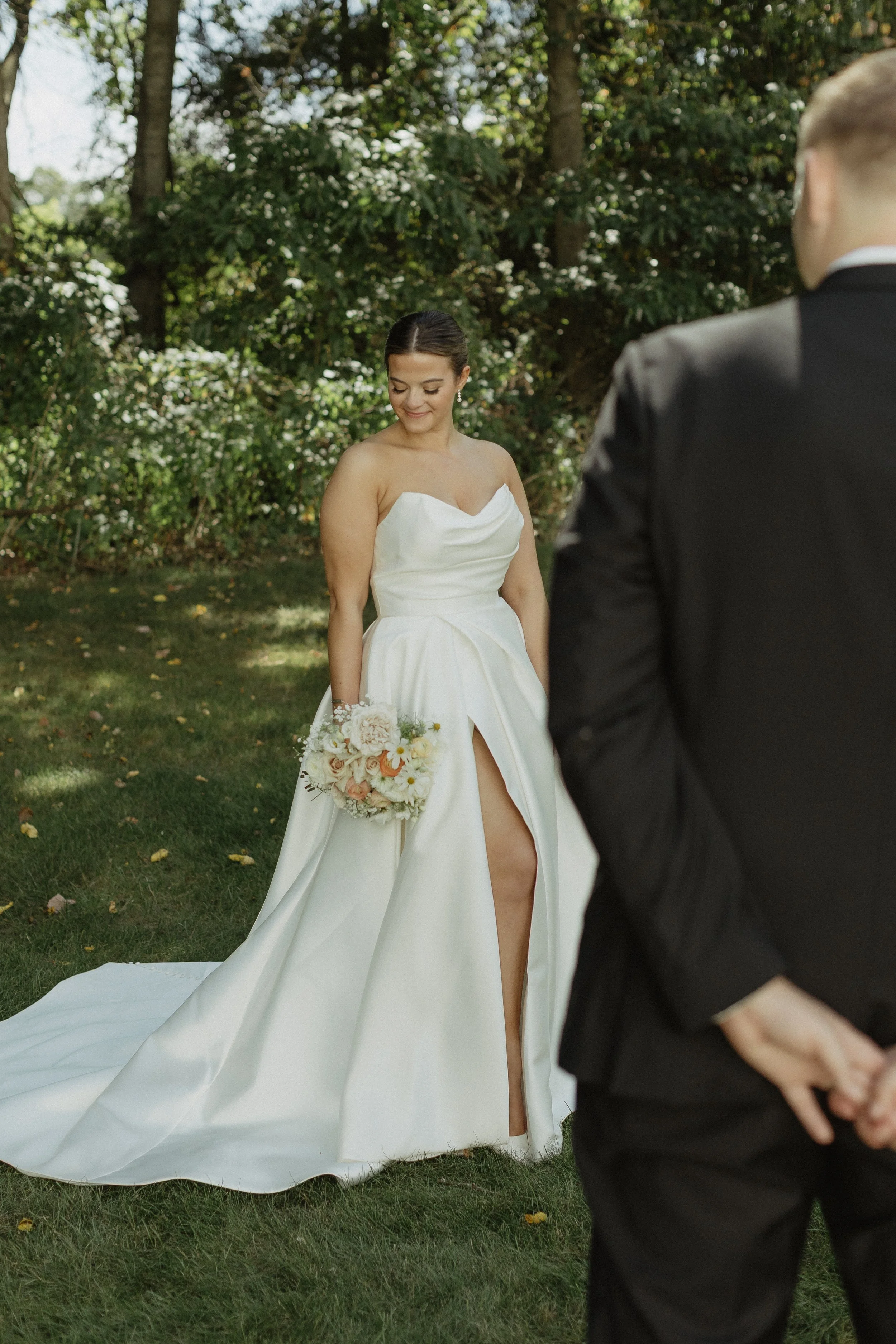 A bride in a satin wedding gown holding a bouquet of flowers, standing outdoors on grass with trees in the background. A groom in a black suit stands in front of her with his hands clasped behind his back.