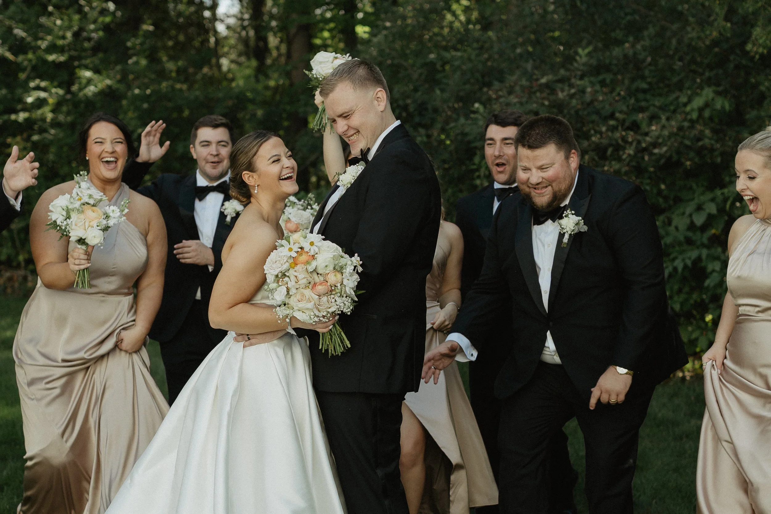 A candid wedding group photo outdoors featuring a bride and groom surrounded by bridesmaids and groomsmen, all smiling and laughing.