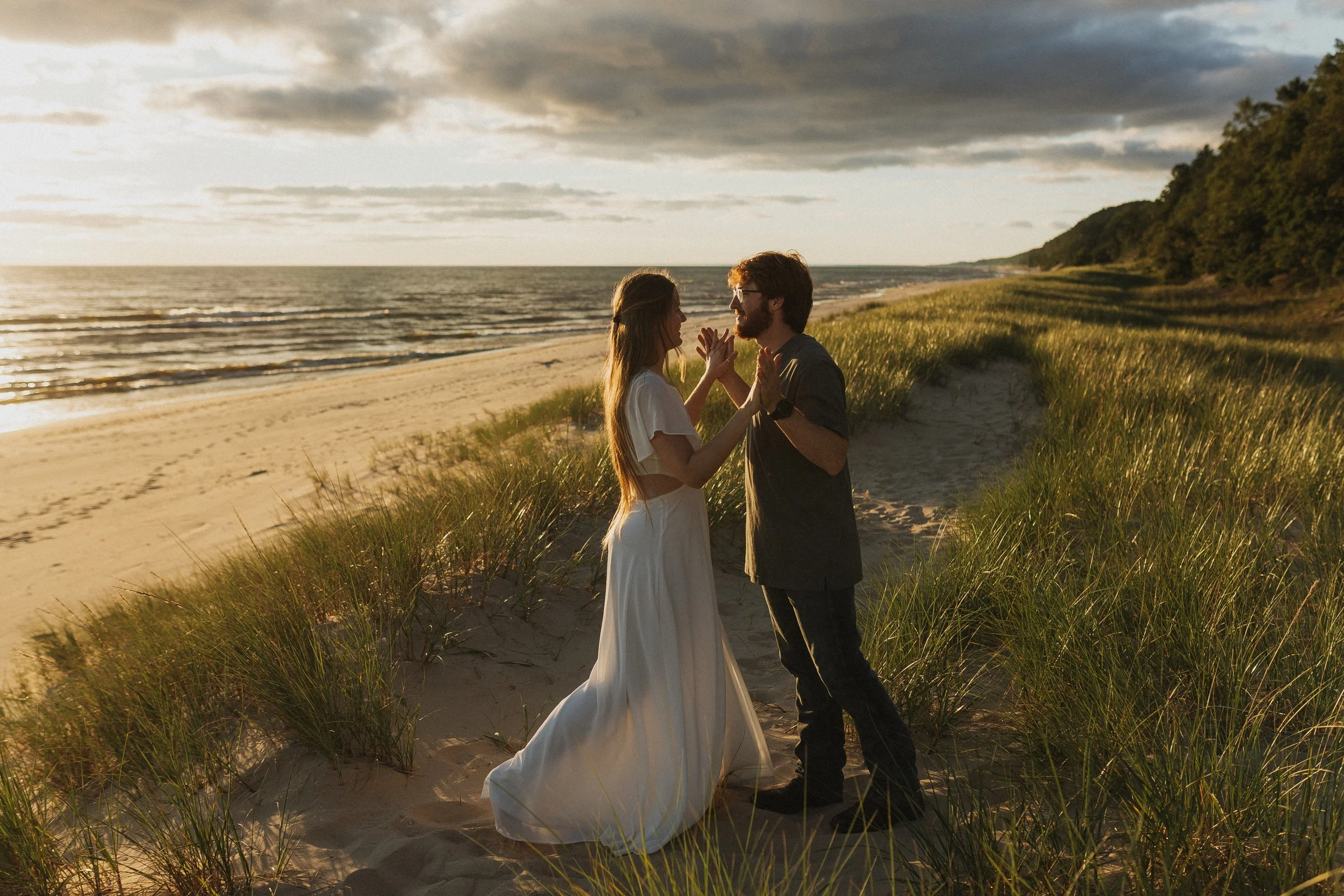 A couple standing on a sandy beach, holding hands and gazing into each other's eyes during sunset, with grassy dunes in the foreground and the ocean in the background.