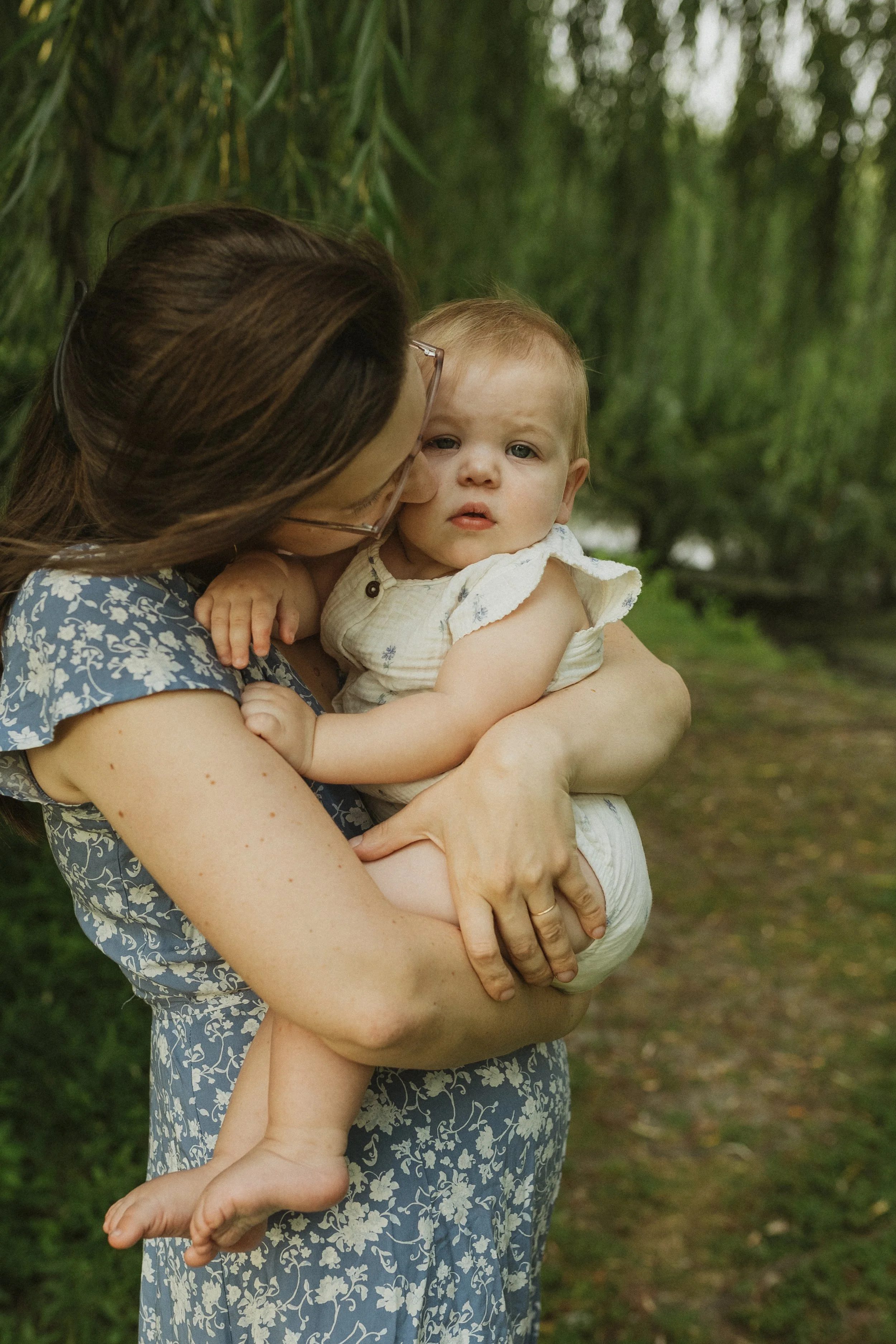 A woman holding a young child outdoors in a green, wooded area. The woman is wearing glasses and a blue dress with white floral patterns. The child, looking at the camera with a slightly serious expression, is dressed in a light-colored sleeveless ou