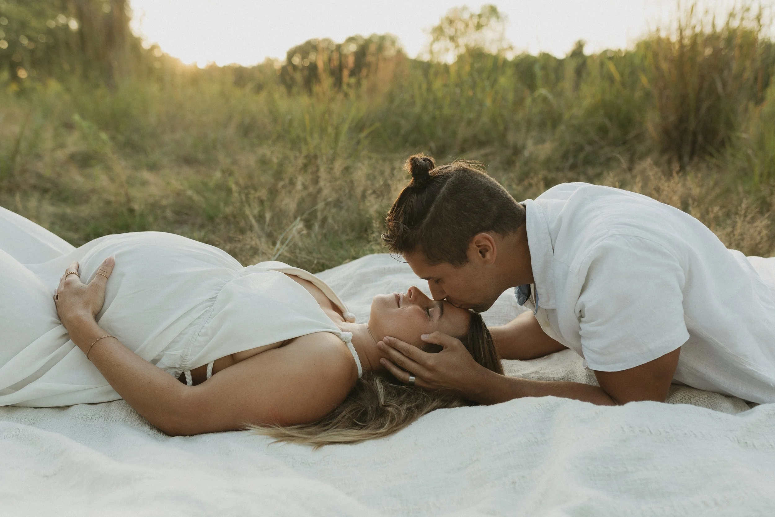 A couple lying on a blanket outdoors, with the man gently holding the woman's face as they kiss, surrounded by nature