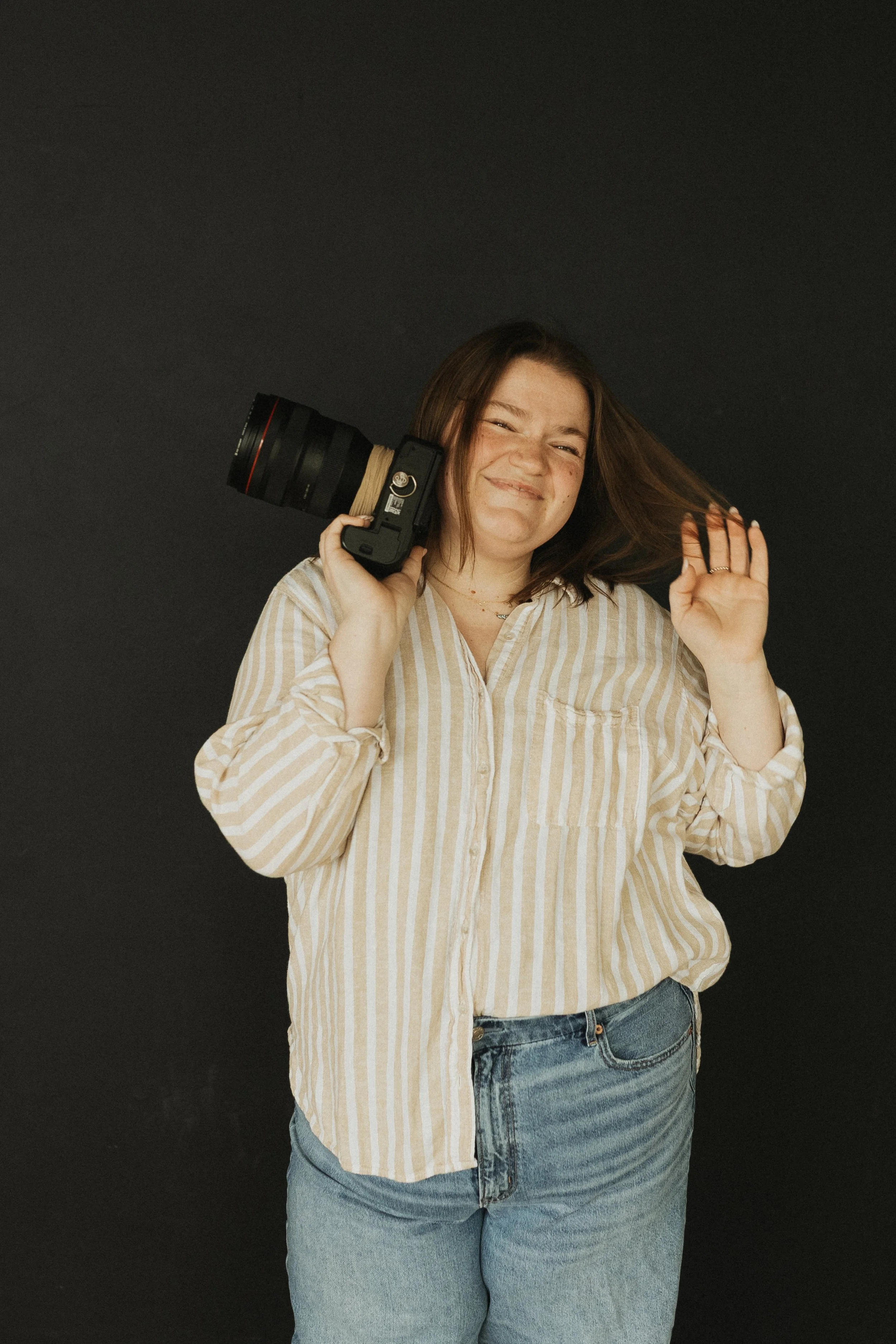 Young woman smiling, holding a professional camera on her shoulder, waving, against a plain dark background.