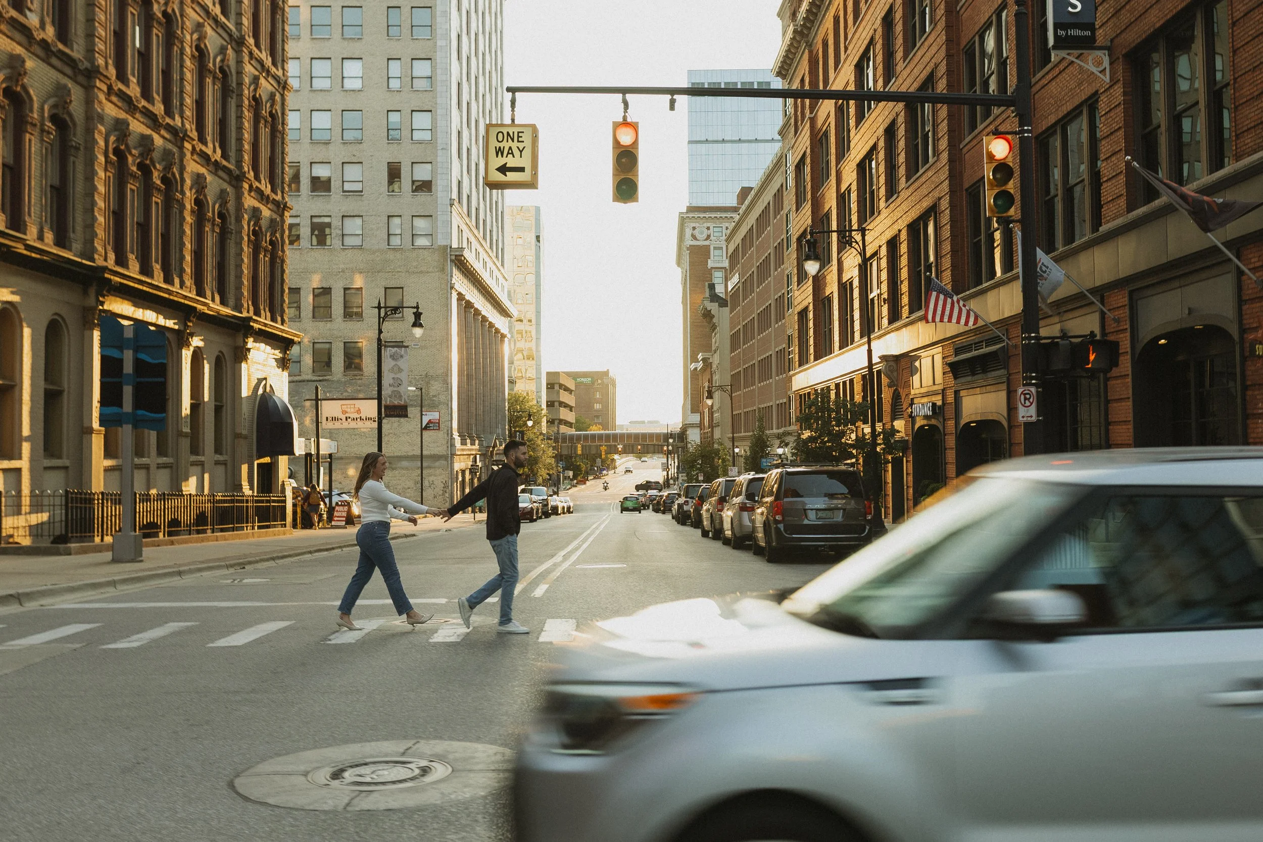 Two people holding hands and crossing a city street at a crosswalk with cars parked along the side, tall buildings, traffic lights, and American flags in the background during daylight.