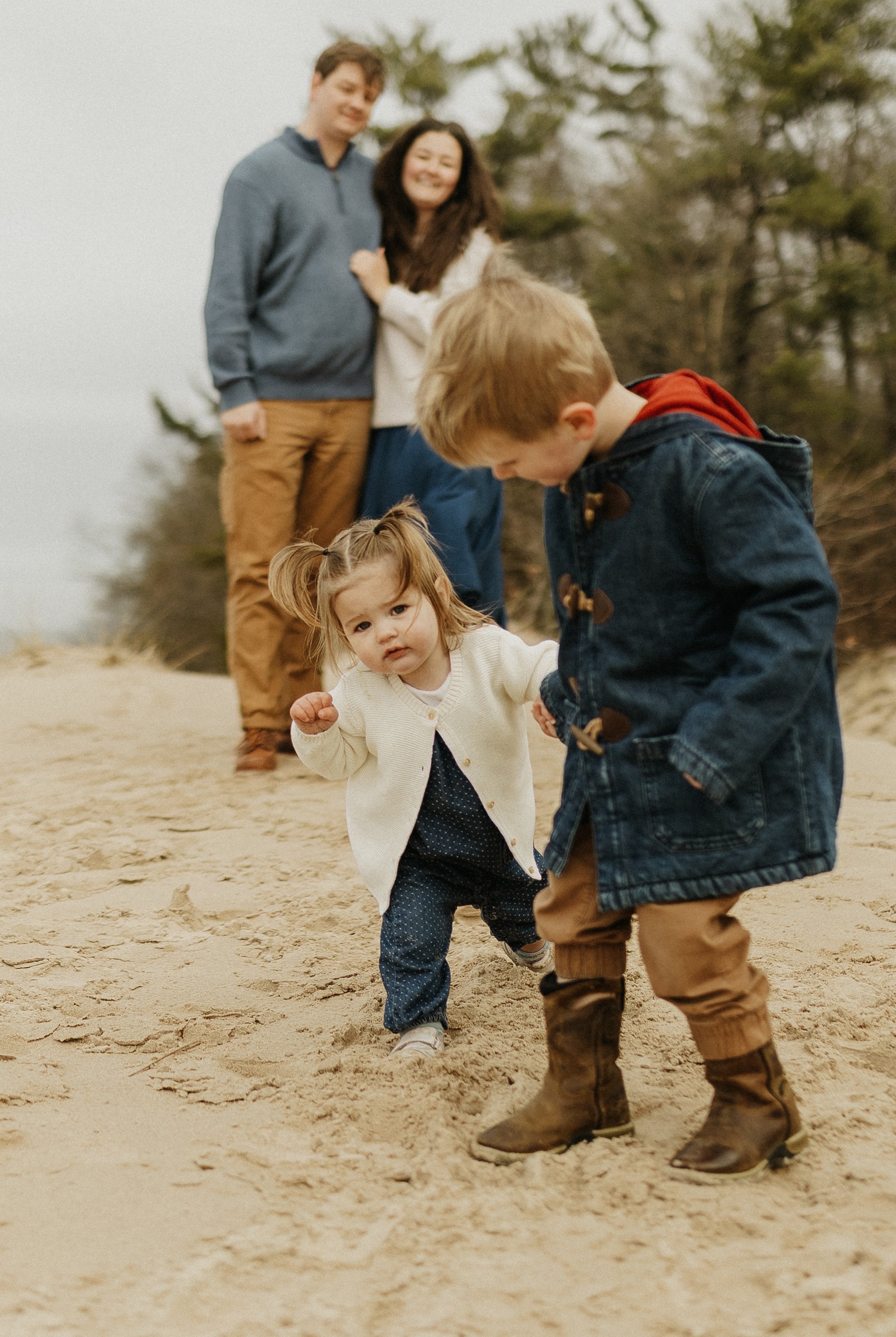 A family of five walking on a sandy beach with trees in the background. Two adults and two children are visible, with a young girl and boy in the foreground, and a man and woman standing behind them.