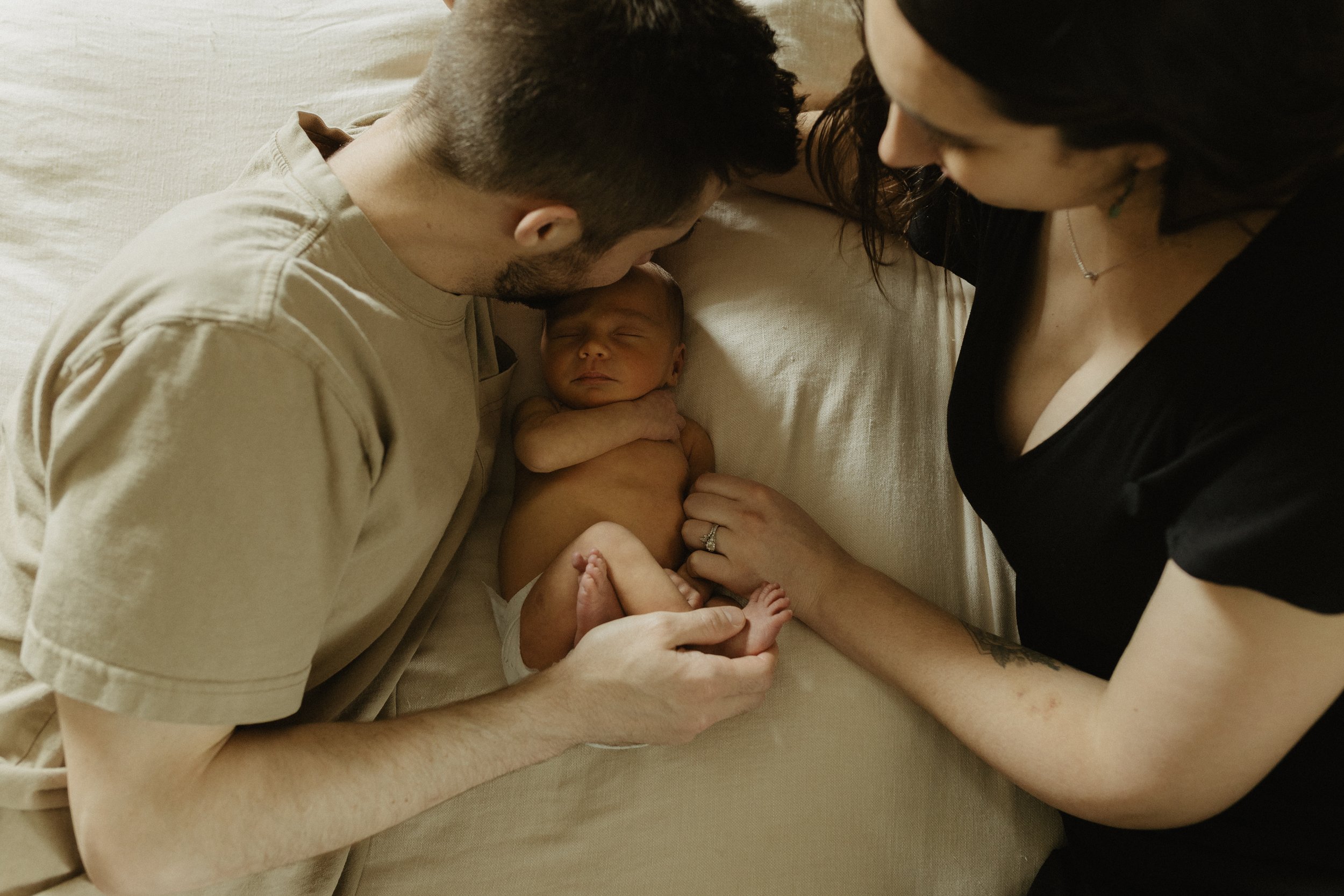 A newborn baby is lying on a beige bed with closed eyes, surrounded by a man and a woman. The man and woman are gently touching the baby and looking at it lovingly, creating a tender family moment.