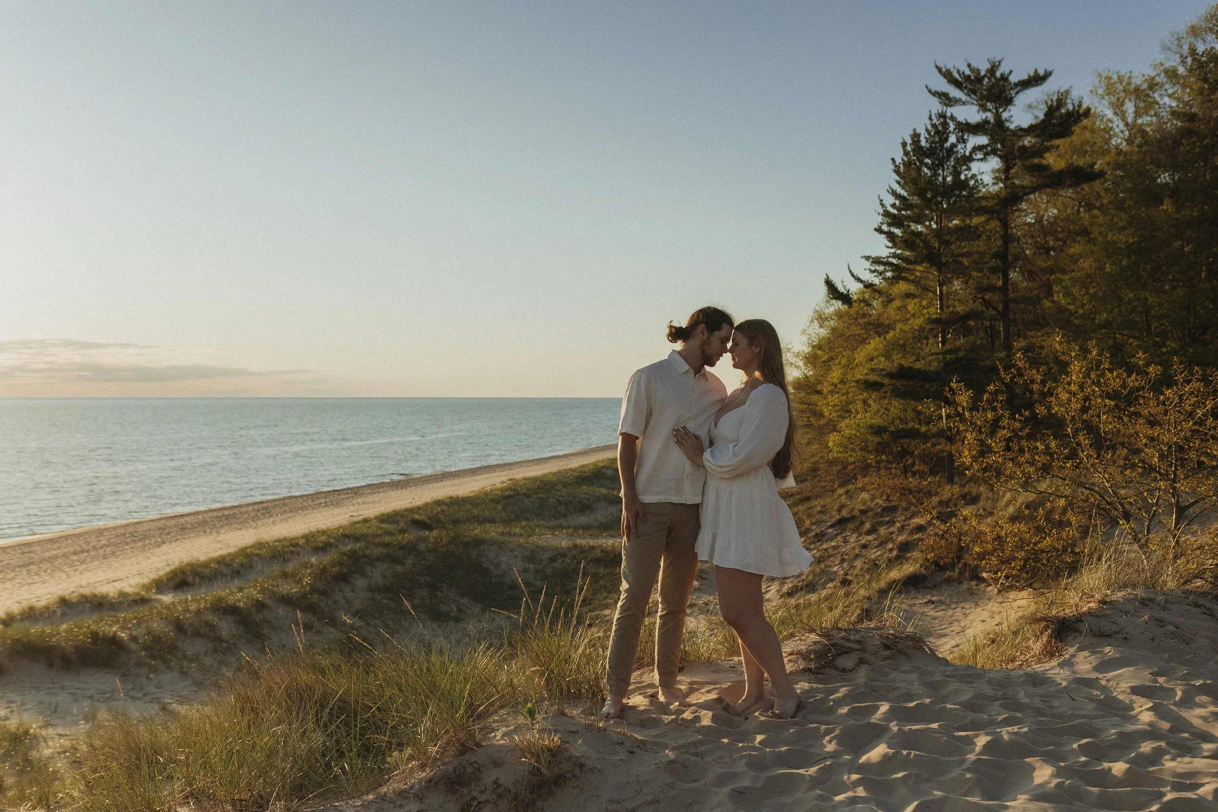 A couple standing close together on a sandy beach at sunset, with the ocean in the background and trees on the right side.