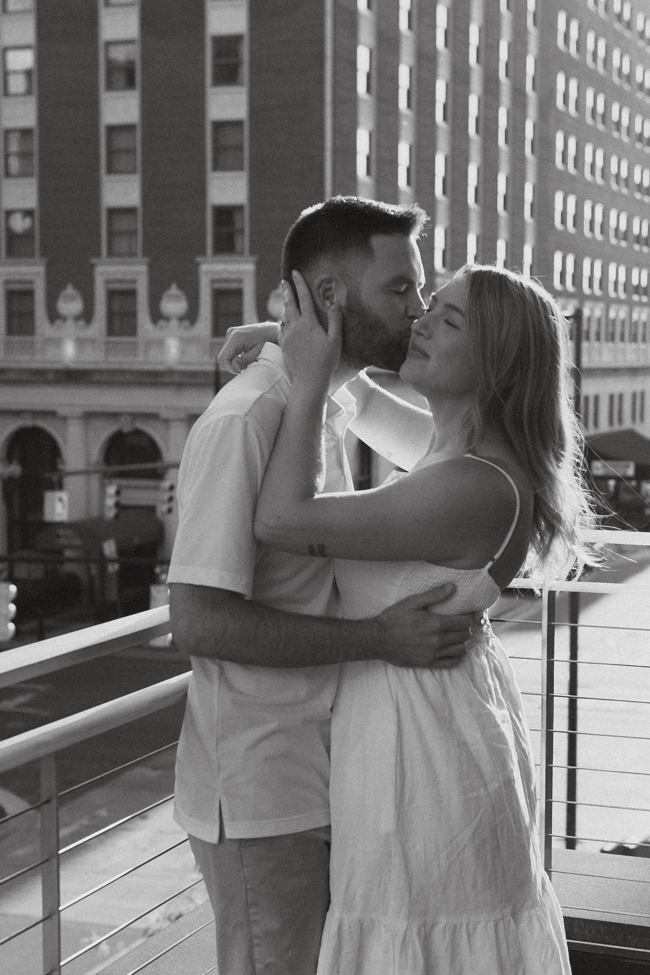 A black-and-white photo of a couple embracing on a balcony, with the man kissing the woman on the forehead, urban buildings in the background.