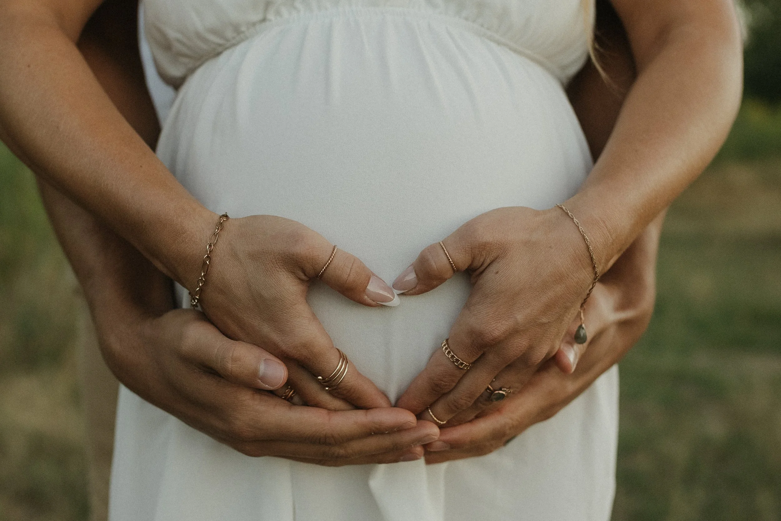 A pregnant woman is holding her belly with both hands, which are decorated with rings and bracelets, forming a heart shape.
