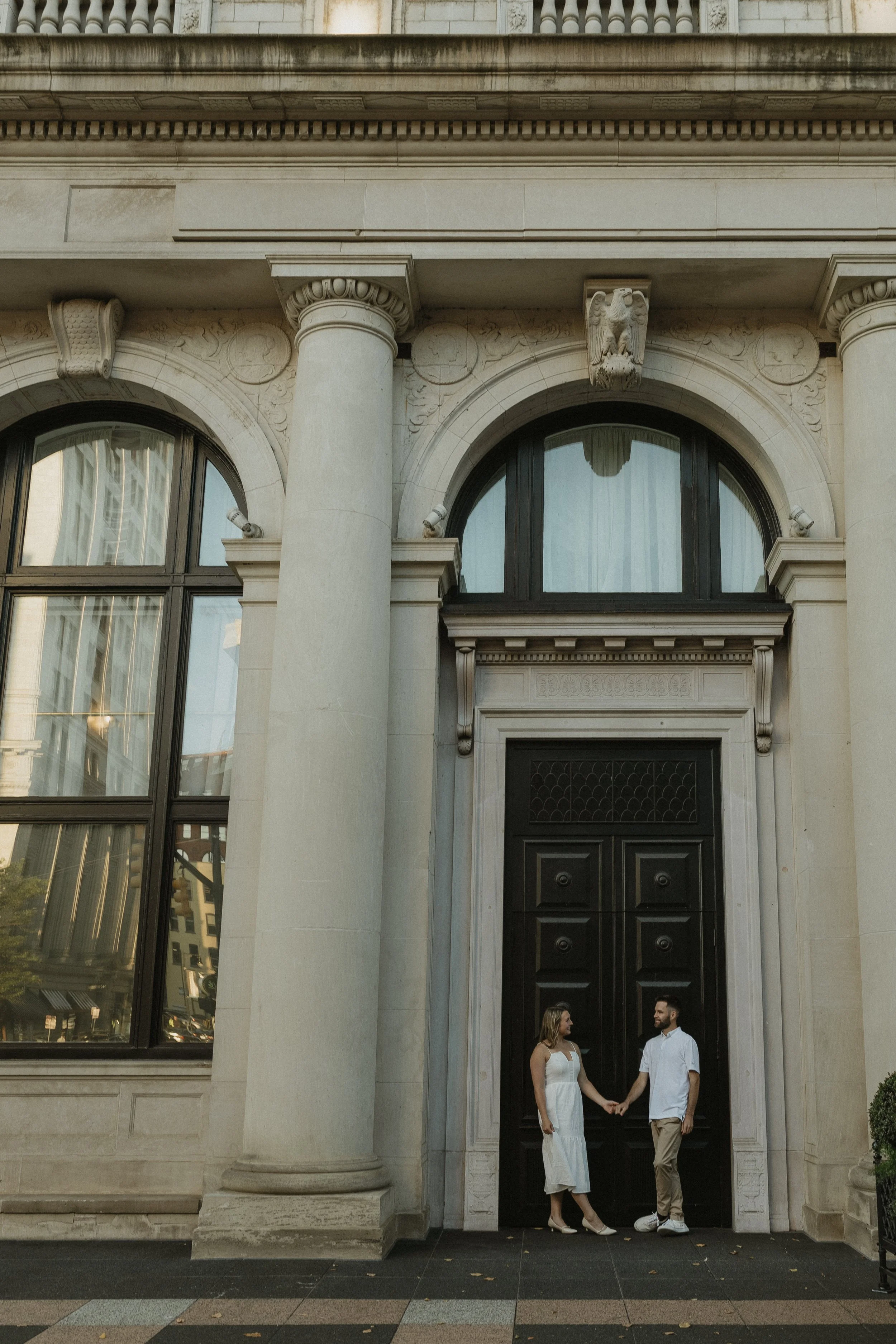 A man and a woman holding hands standing in front of a large black door on a classical building with ornate architectural details.